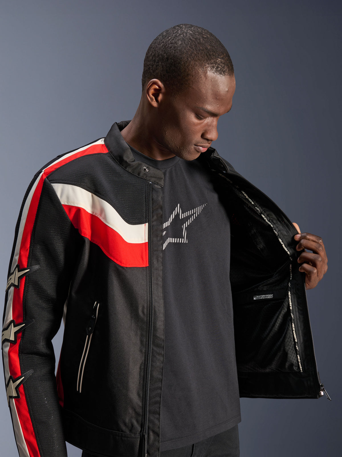 Alpinestars T-Dyno Air Jacket, svart, klarröd och ecru, manlig modell som öppnar jackan för att visa det inre fodret och det vattentäta facket, vintageinspirerad textiljacka med 3D-meshpaneler och stjärnlogotyper, modern motorcykelutrustning
