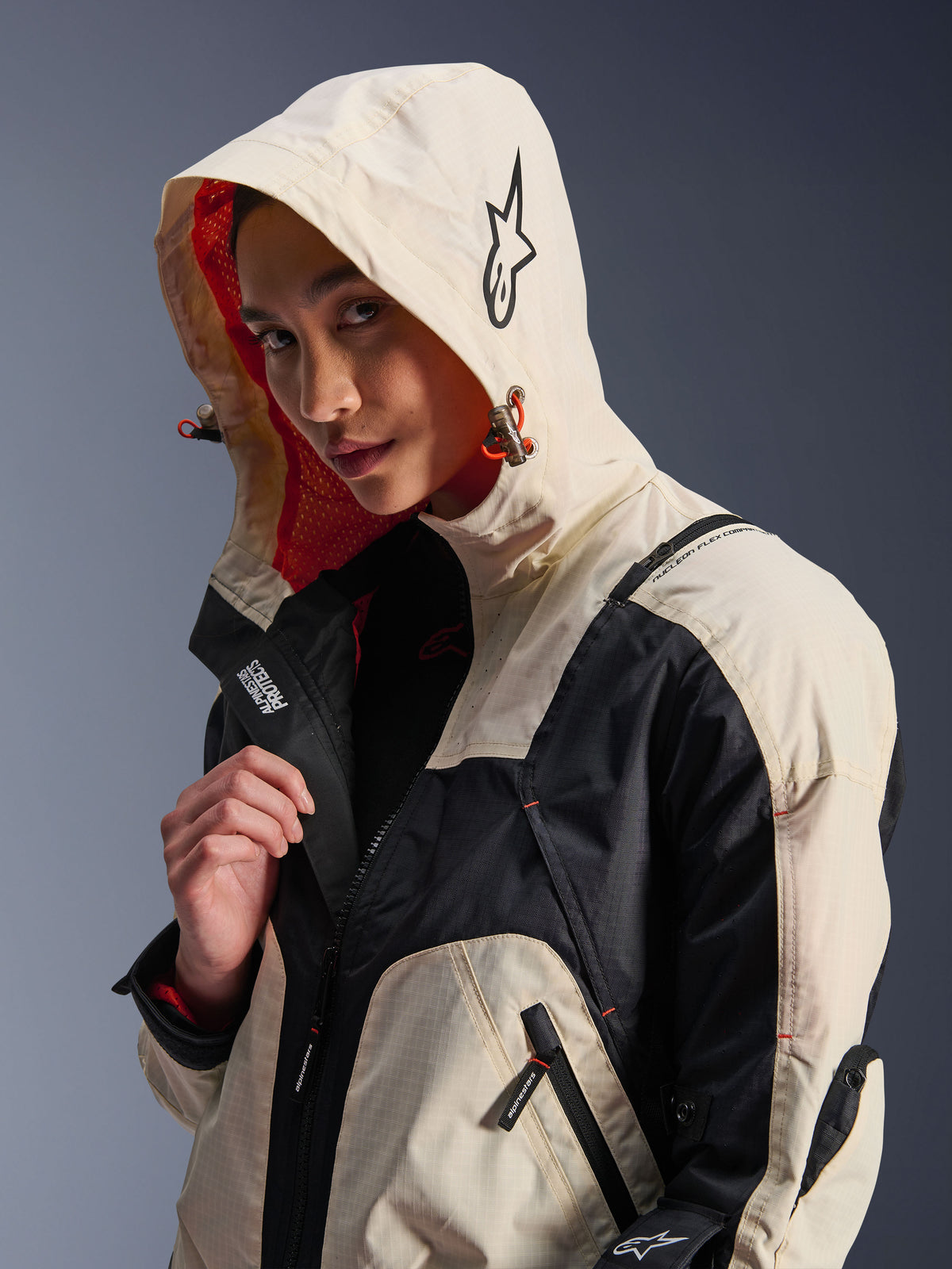 Alpinestars Molly Air-jacka i Black Almond, bärs av en kvinnlig modell med huvan uppfälld, krämvit och svart färgblockering, livfullt orange meshfoder, tekniska urbana designdetaljer med stjärnlogotyp och dragkedjor