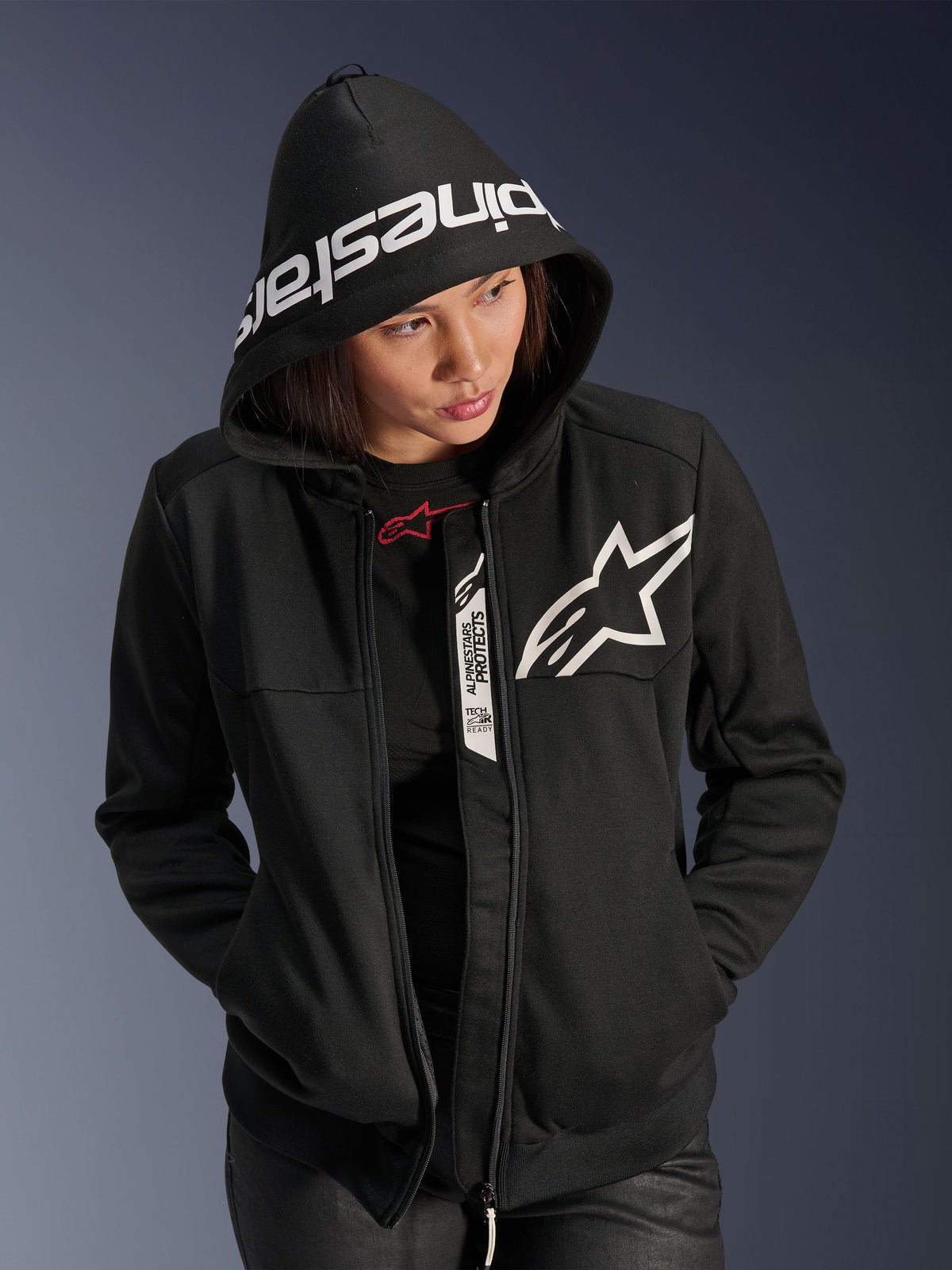 Alpinestars Stella Chrome V2 Sport Hoodie för dam, svart och vit, motorcykeltröja med en vit stjärnlogotyp på bröstet och Alpinestars-märkning på huvans kant, kvinna med huvan uppfälld, avslappnad urban sportstil med Tech-Air Ready-detaljer