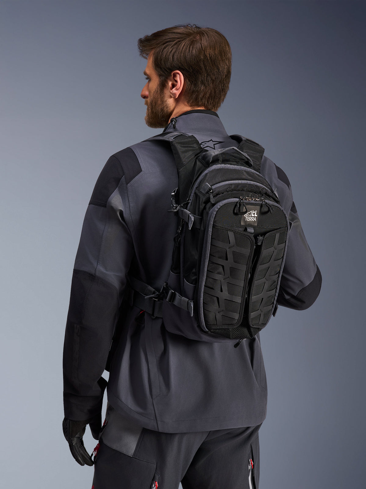 Alpinestars AT-15 ryggsäck, äventyrsryggsäck för motorcykel, svart och mörkgrå, bärs av en förare i en grå jacka, teknisk design med MOLLE-system och 'TERRA'-logotyp, ergonomisk terrängutrustning