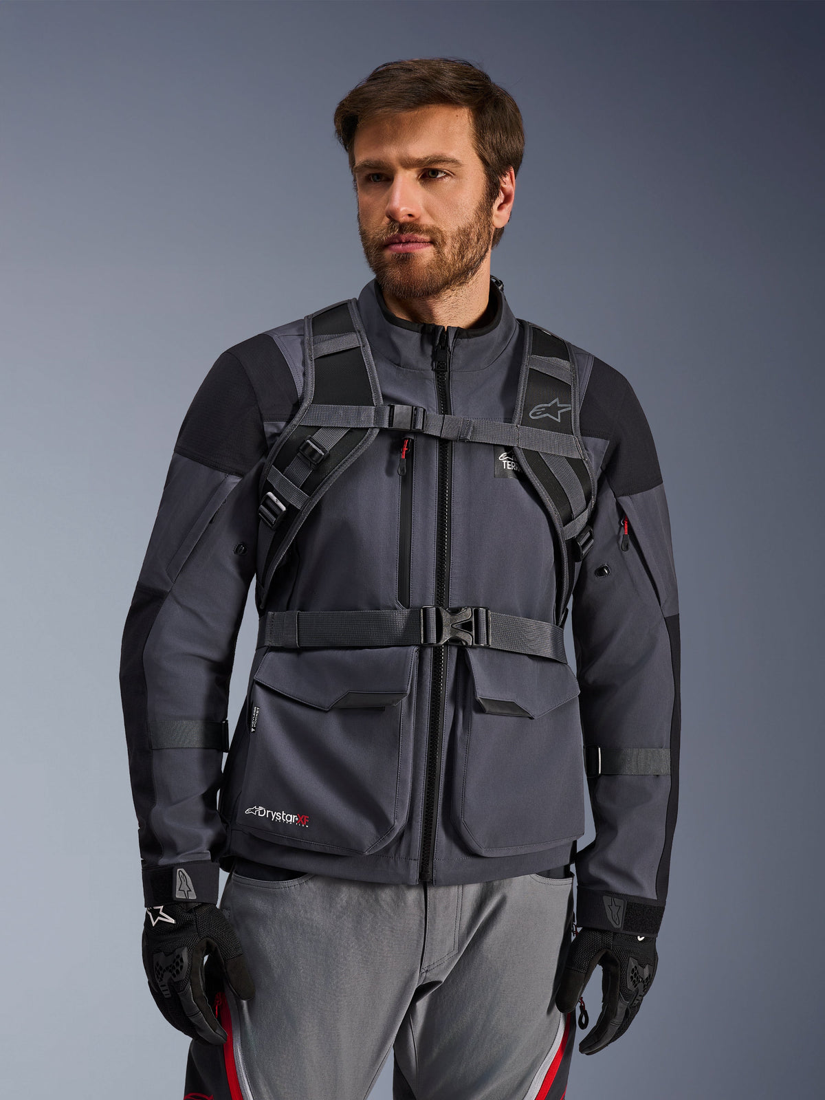 Alpinestars AT-15 Back Pack bärsystem, Black Deep Gray, framifrån buren av en förare över en grå motorcykeljacka, med magnetiskt bröstlås och säkert midjebälte, matchad med svarta handskar för äventyrsmotorcykelkörning