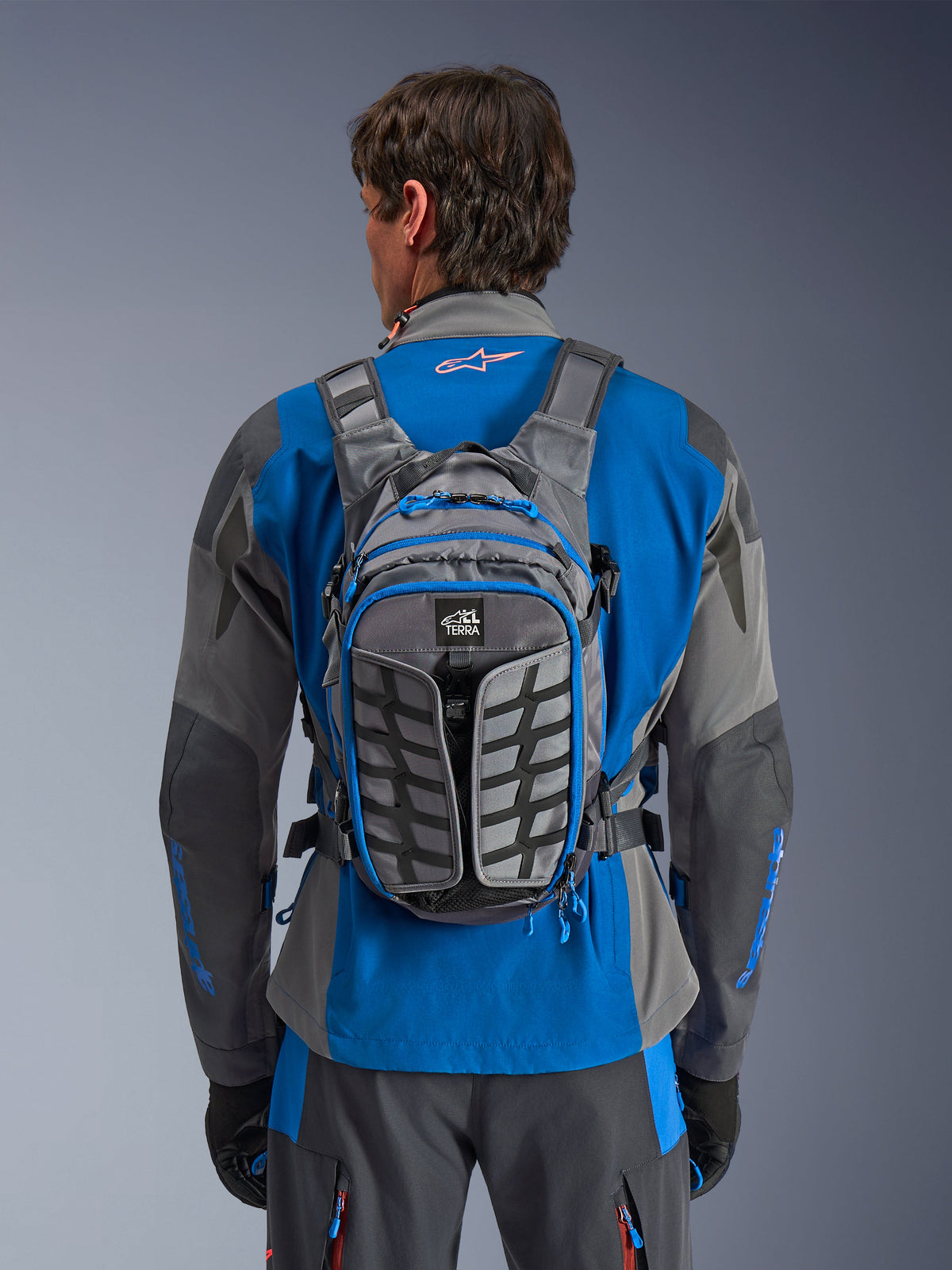 Alpinestars AT-15 Back Pack, äventyrsryggsäck för motorcykel, mörkgrå och djupgrå, bärs av en förare över en blå och grå jacka, med All Terra-branding, ergonomisk strukturell design, justerbara remmar och blå dragkedjsdetaljer.