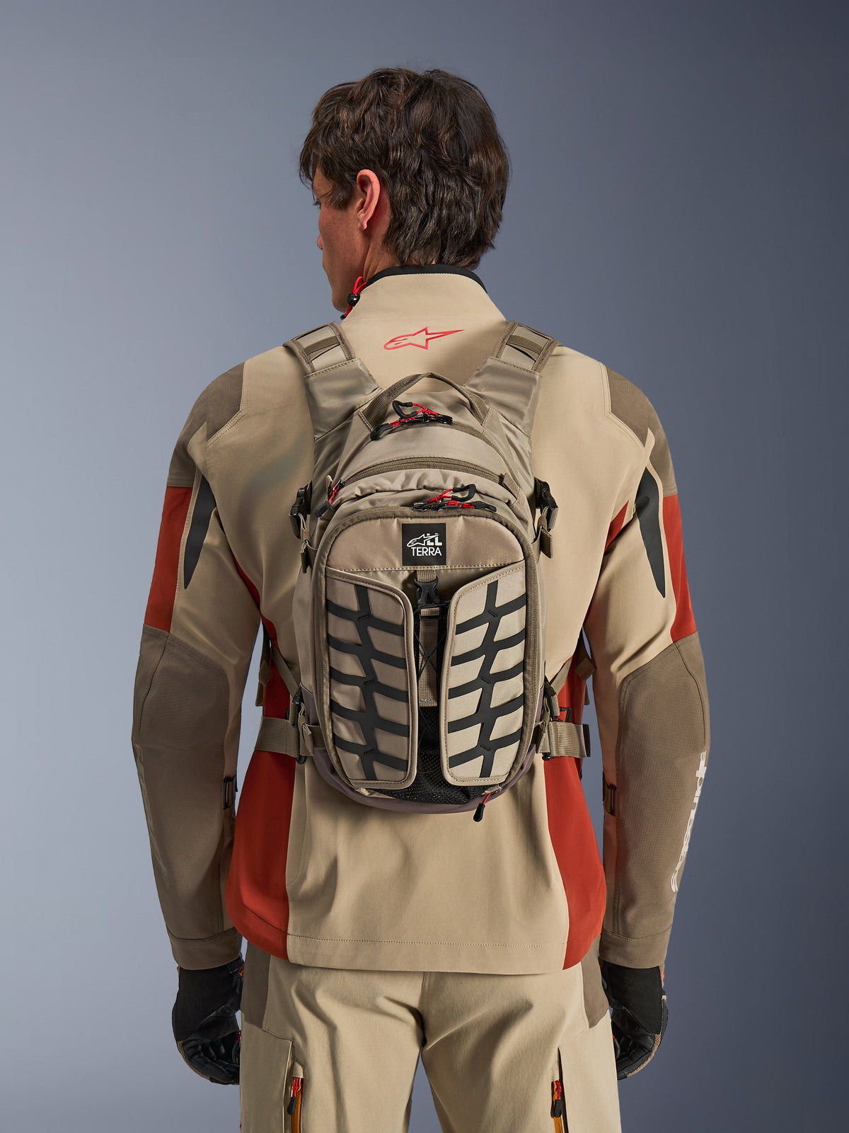 Alpinestars AT-15 Back Pack, Aluminum Walnut, teknisk äventyrsryggsäck för motorcykel buren av en förare i en beige och röd jacka, med däckmönster, TERRA-logotyp, röda dragtappar och justerbara remmar.
