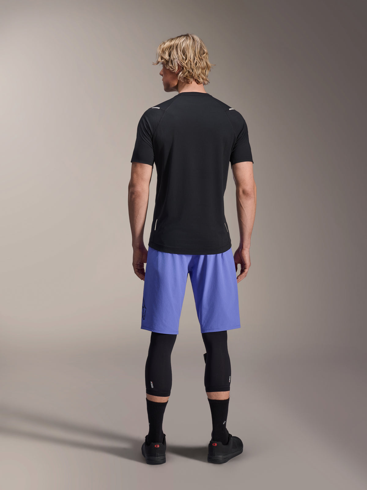 Male rider wearing Alpinestars A-Aria Lite Shorts, MTB Shorts, Baja Blue, back view, styled with a black short-sleeve jersey, black knee guards, and black cycling shoes, performance mountain biking apparel