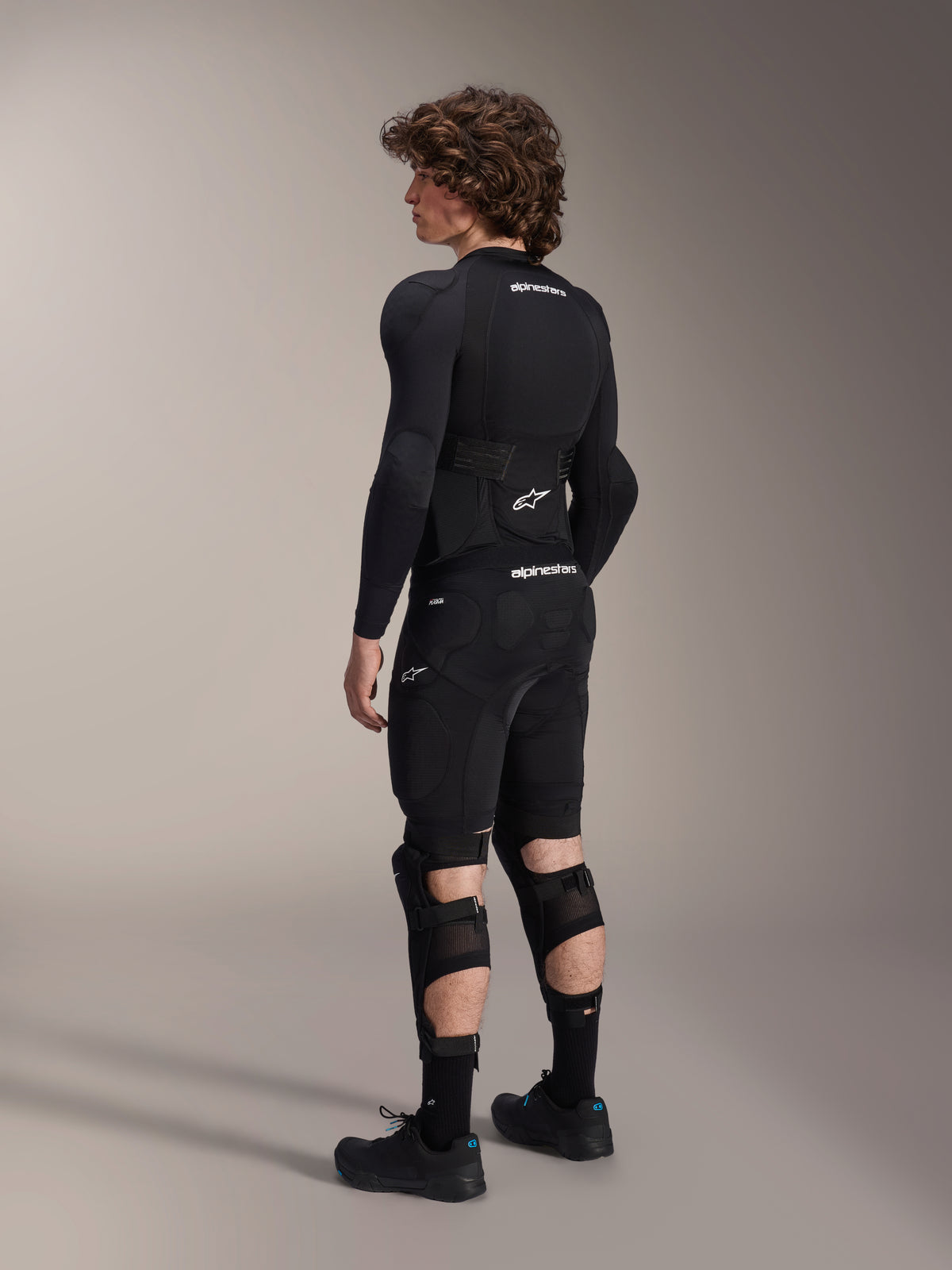 Alpinestars A-Impact Plasma Pro knä- och skenbensskydd sedda bakifrån, svarta och vita, visar det trippla remsystemet och ventilerande meshpaneler, bärs med en komplett uppsättning svart skyddsutrustning för mountainbike inklusive långärmat underställ och vadderade shorts