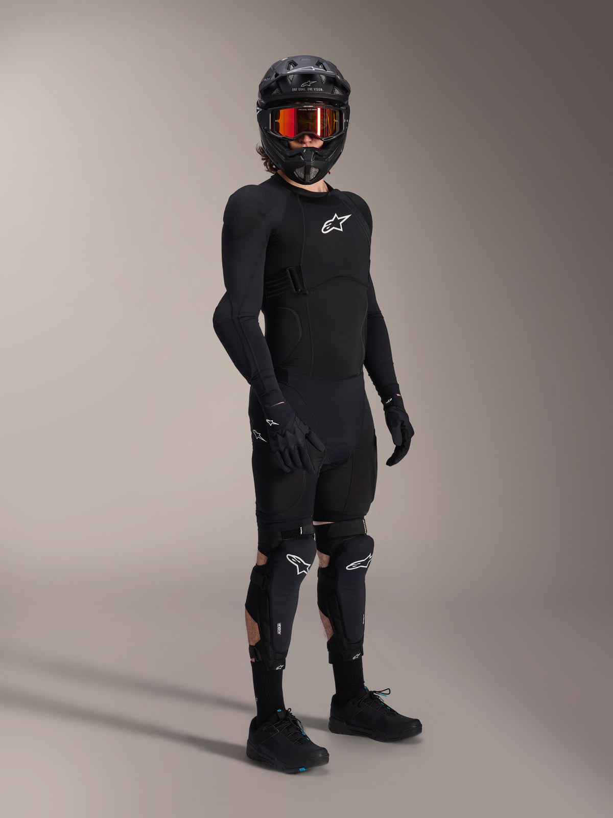 Alpinestars A-Impact Plasma Pro knä- och skenbensskydd, svarta och vita, bärs av en cyklist med en komplett skyddsutrustning för mountainbike, inklusive en svart långärmad skyddströja, svarta handskar och en fullface-hjälm med orangea goggles.