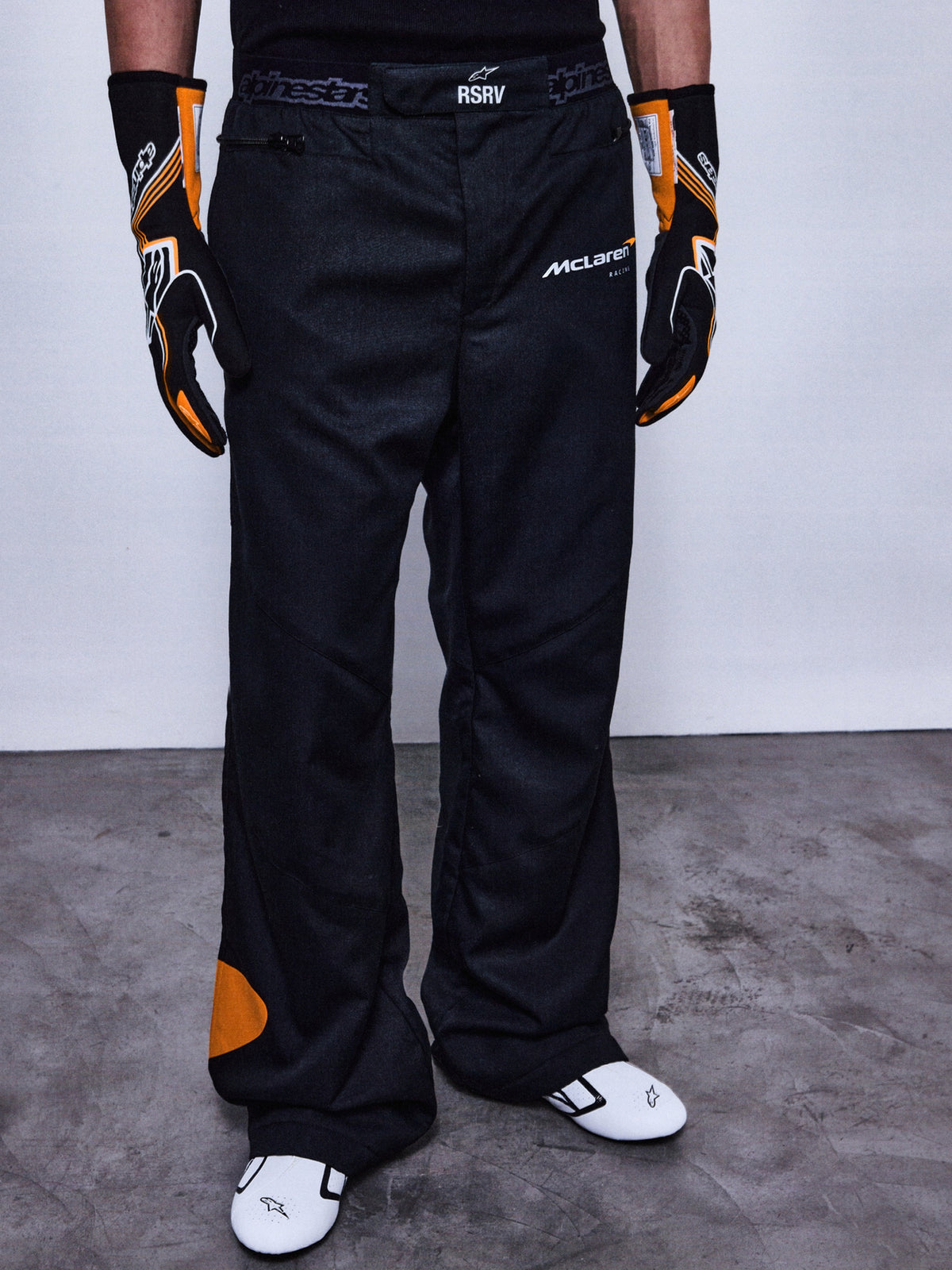 Alpinestars RSRV Arena Suit Pants, svarta racingbyxor med McLaren-logotyp, orangea detaljer på sidan och knä, person iklädd med svarta och orangea handskar samt vita racingskor
