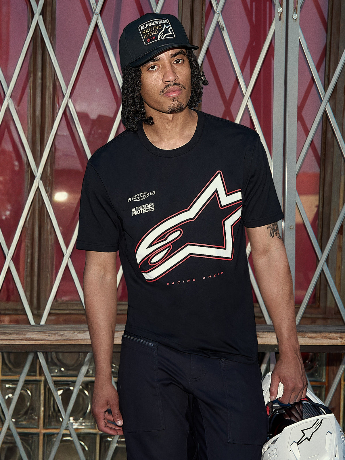 Alpinestars Authority CSF Tee, kortärmad t-shirt, svart, stor vit och röd Astars-konturlogotyp med 'Alpinestars Protects'-grafik, livsstilsbild på en man i svart trucker-keps som håller en vit hjälm, casual motorsport-streetwear