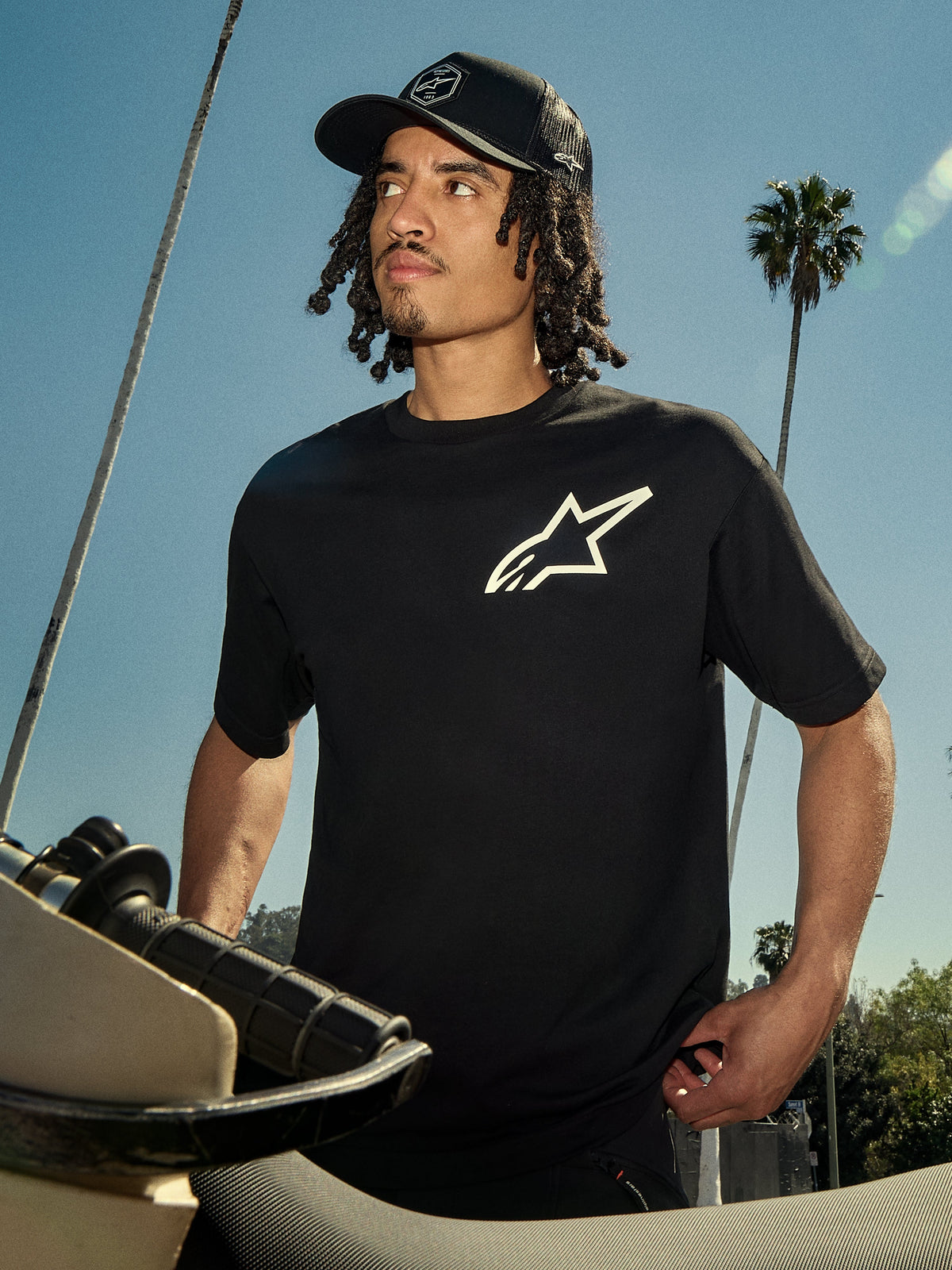 Alpinestars One Oversized CSF Tee, kortärmad t-shirt, svart med vit Astars-logotyp, bärs av en man i en svart truckerkeps, utomhus livsstilsmiljö med palmer och en motorcykel, avslappnad motorsport-streetwear