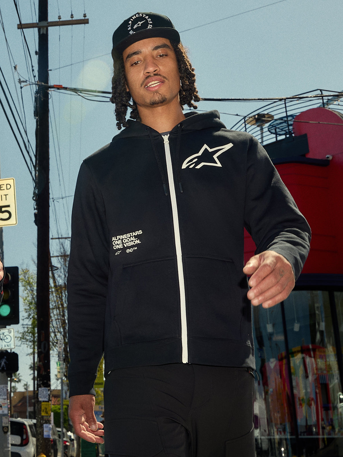Alpinestars Transverse Hoodie, svart huvtröja i fleece med dragkedja, vit dragkedja och vit Astars-logotyp på bröstet, "One Goal One Vision"-texttryck, avslappnad streetwear-stil, bärs av en man i en urban utomhusmiljö
