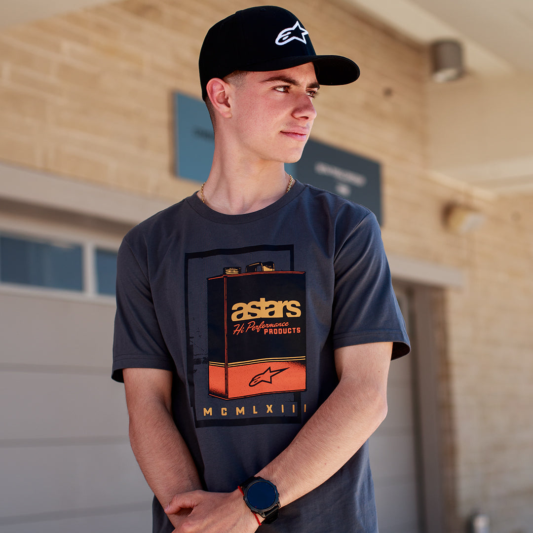 Alpinestars Galun CSF Tee, kortärmad t-shirt, mörkgrå, vintage bensindunk-grafik fram med 'astars'-märkning och romerska siffror 'MCMLXIII', buren av en man med en svart keps med logotyp, avslappnad motorsport-livsstil