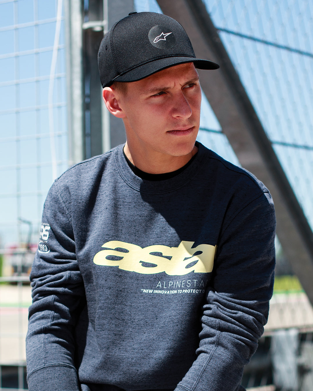 Fabio Quartararo bär Alpinestars Apex Keps i svart, keps med strukturerad krona, böjd skärm och cirkulär stjärnlogotyp, kombinerad med en mörkgrå melerad sweatshirt med gul märkning, avslappnad motorsportinspirerad livsstil.