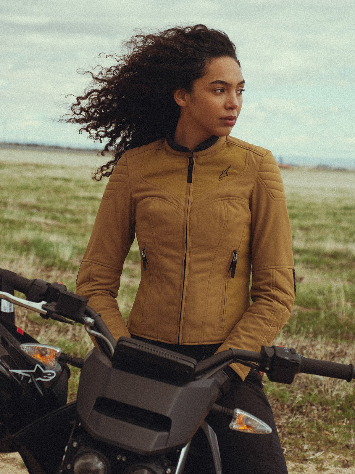 Alpinestars Isla WR damjacka i Utility Brown Light Gold, bärs av en kvinna som sitter på en motorcykel på ett fält utomhus, med quiltade detaljer på axlarna, en slim-fit-silhuett och urban modern stil.