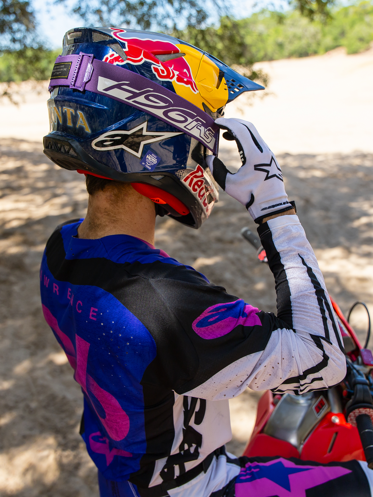 Motocrossföraren Hunter Lawrence bär Limited Edition HUNTA96 XXV Supertech Vision Goggles, lila rem med 'H96' och gula sömmar, blå Red Bull-hjälm, lila och vit tröja med svarta detaljer, vita handskar, justerar glasögonen i en sandig terrängmiljö