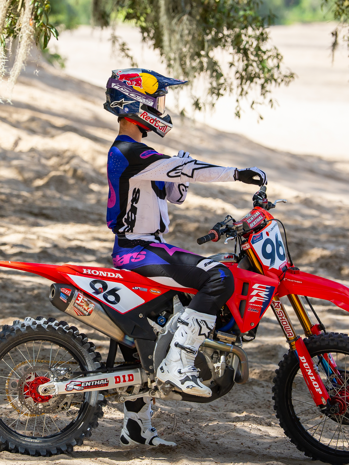 Motocrossförare som bär Limited Edition HUNTA96 XXV Supertech Vision Goggles i Purple White Mirror Silver, matchande lila och vit racingtröja med vita stövlar, sittande på en röd Honda motocrosscykel med nummer 96, utomhusmiljö för terrängkörning på en sandig bana.