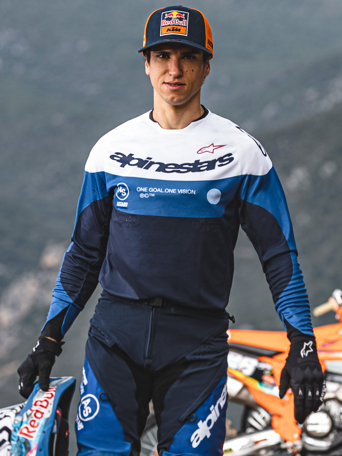 Motocrossförare i Alpinestars Pro-Dura-tröja och byxor i marinblått, klarblått och vitt, med 'One Goal One Vision'-profilering, svarta handskar, håller i en racinghjälm, professionell offroad-racingutrustning