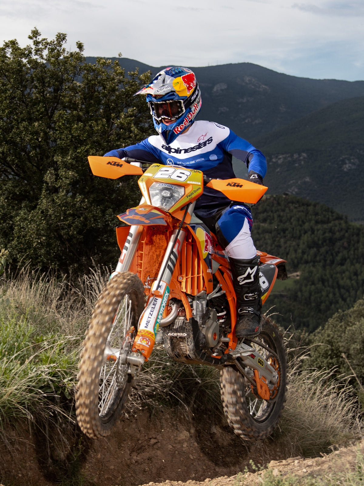 Motocrossförare i action på en orange KTM-dirtbike, iklädd Alpinestars Pro-Dura-utrustning i marinblått, klarblått och vitt. Professionell offroad-outfit med blå och vit tröja och matchande byxor, buren med en Red Bull-hjälm och svarta stövlar på en bergsstig.