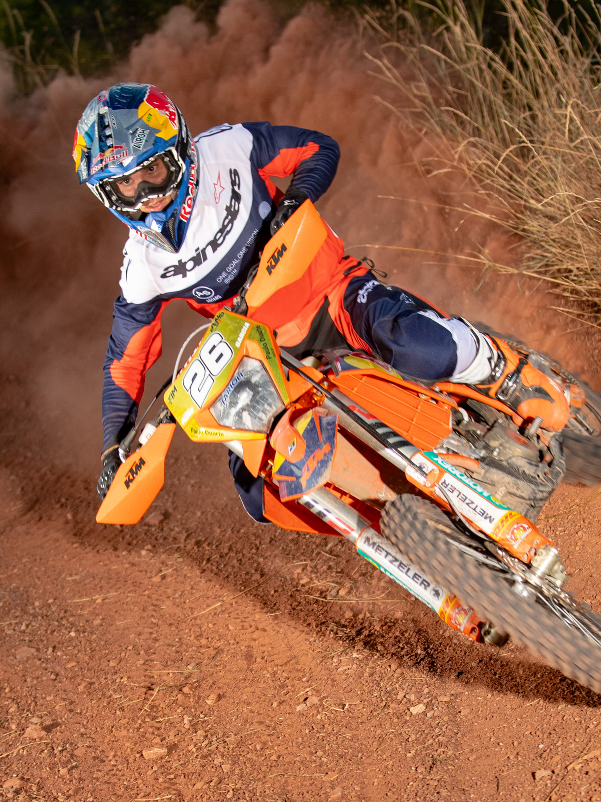 Motocrossförare i en kurva på en orange motorcykel, bär Alpinestars Pro-Dura tröja och byxor i marinblått, orange och vitt, matchande utrustning för terrängkörning, Red Bull-märkt hjälm och vita stövlar, actionbild på dammig bana.