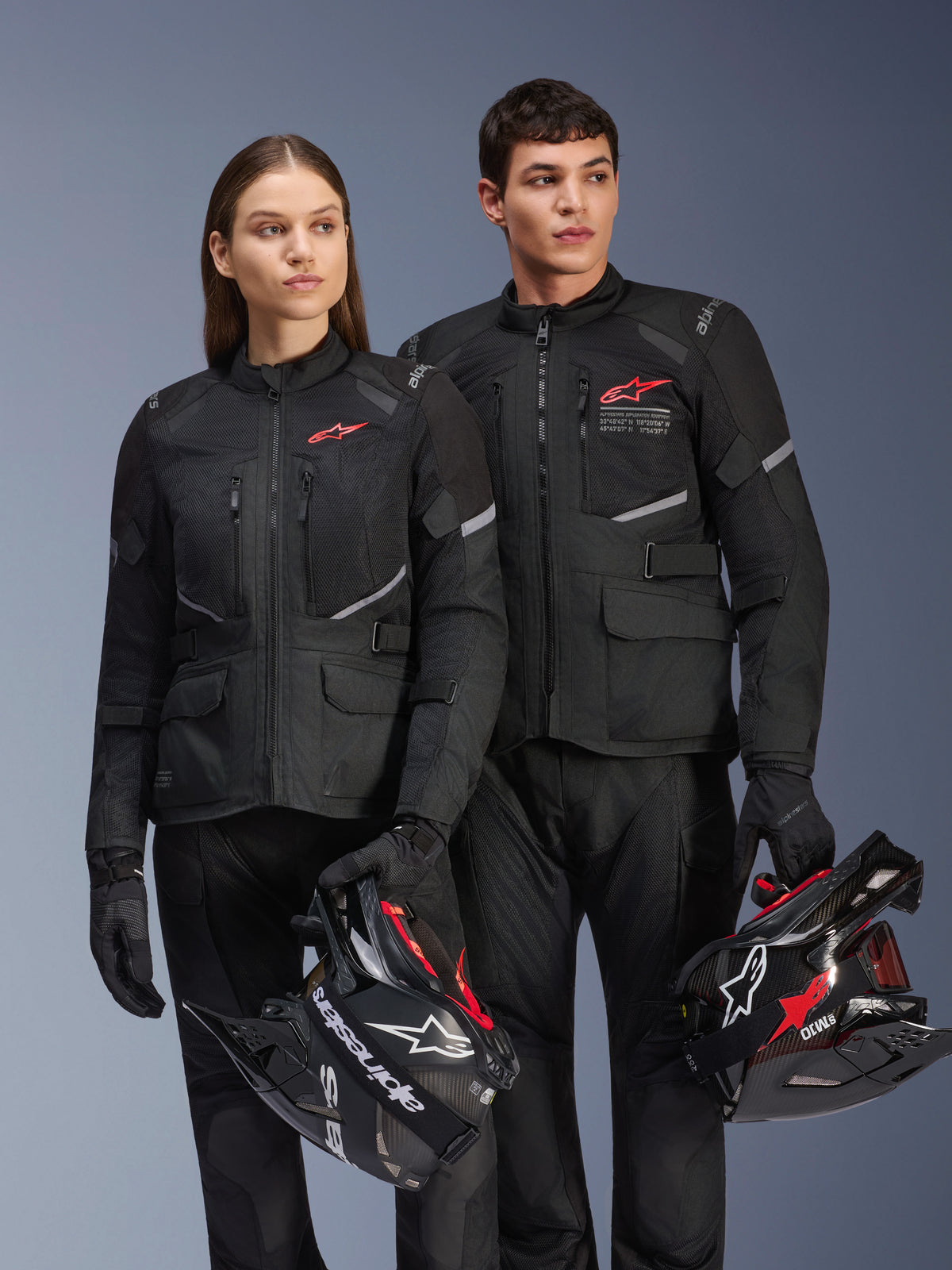 Man och kvinna iförda Alpinestars Andes Air Drystar®-jackor, svart adventure touring-utrustning med meshpaneler och reflexdetaljer, matchande svarta byxor och handskar, håller i motocrosshjälmar, studiobild av professionella motorcykelkläder