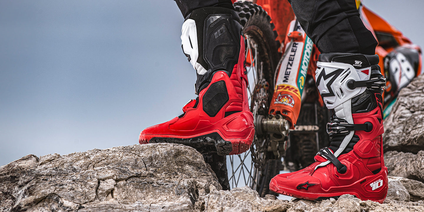 Red, black and white Tech 10 enduro boots on a rocky ridge next to a KTM motorbike