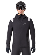 Alpinestars A-Dura regnjacka, MTB-regnjacka, svart med vita axelpaneler, bärs med svart MTB-hjälm, solglasögon och handskar, vattentät design med huva och dragkedja fram, sportig friluftsutrustning för cykling