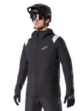 Alpinestars A-Dura regnjacka, regnjacka för mountainbike, svart med ljusgrå axeldetaljer, bärs av en cyklist med svart MTB-hjälm och solglasögon, vit logotyp på bröstet, vattentät konstruktion, matchad med svarta byxor och handskar för terrängcykling