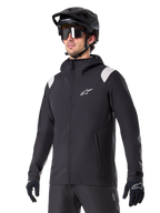 Alpinestars A-Dura regnjacka, regnjacka för mountainbike, svart med ljusgrå axeldetaljer, bärs av en cyklist med svart MTB-hjälm och solglasögon, vit logotyp på bröstet, vattentät konstruktion, matchad med svarta byxor och handskar för terrängcykling