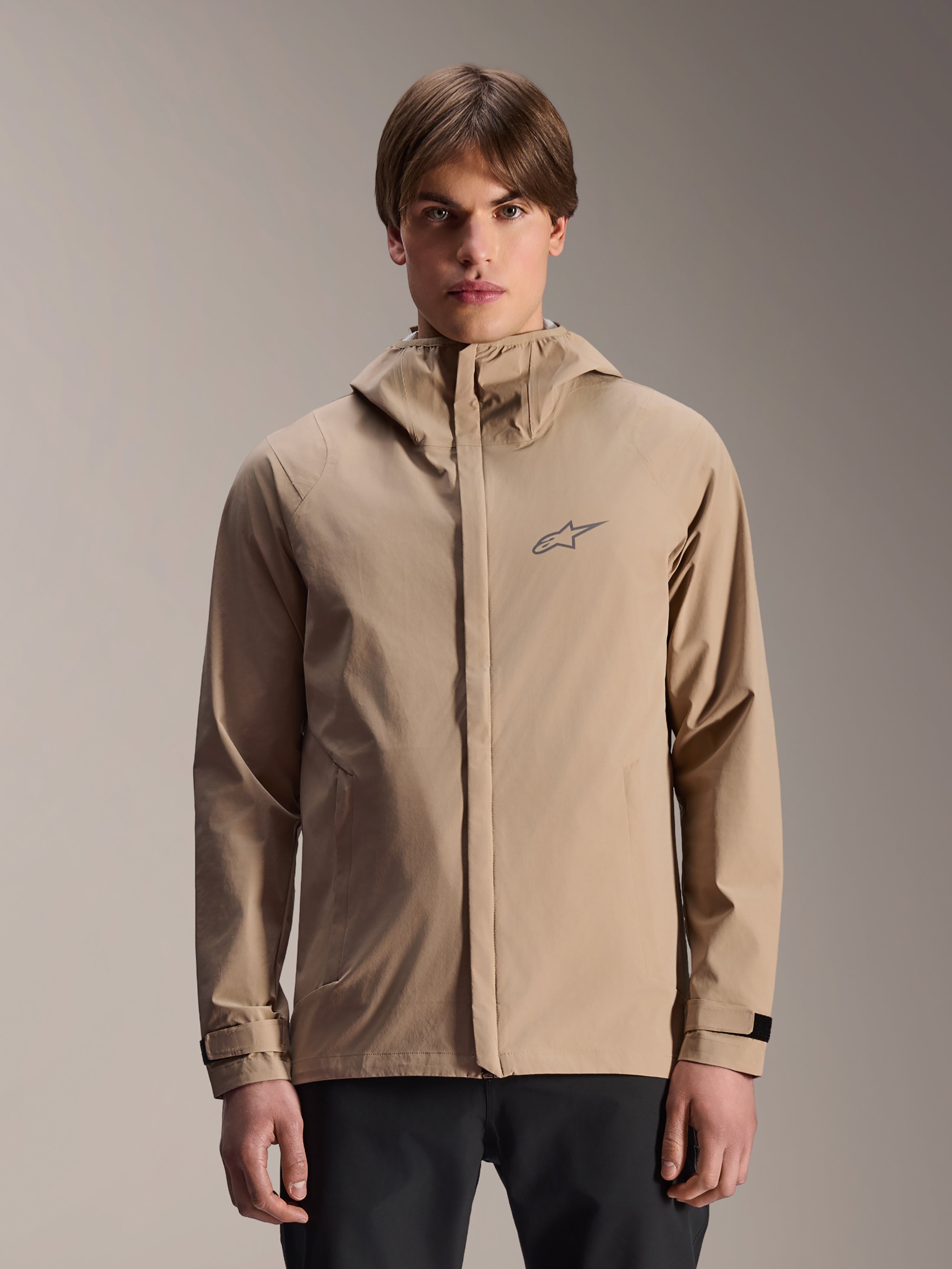 Alpinestars A-Dura Rain Jacket, ljusbrun regnjacka med huva för mountainbike, bärs av en modell, med en liten logotyp på bröstet, justerbara manschetter och dold dragkedja fram, vattentät cykelutrustning