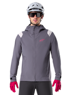 Alpinestars A-Dura regnjacka, MTB-regnjacka, mörk titangrå, design med huva, rosa Alpinestars-logotyp och ljusgrå axelpaneler, bärs med vit MTB-hjälm och starkt rosa handskar, vattentät utrustning för terrängcykling