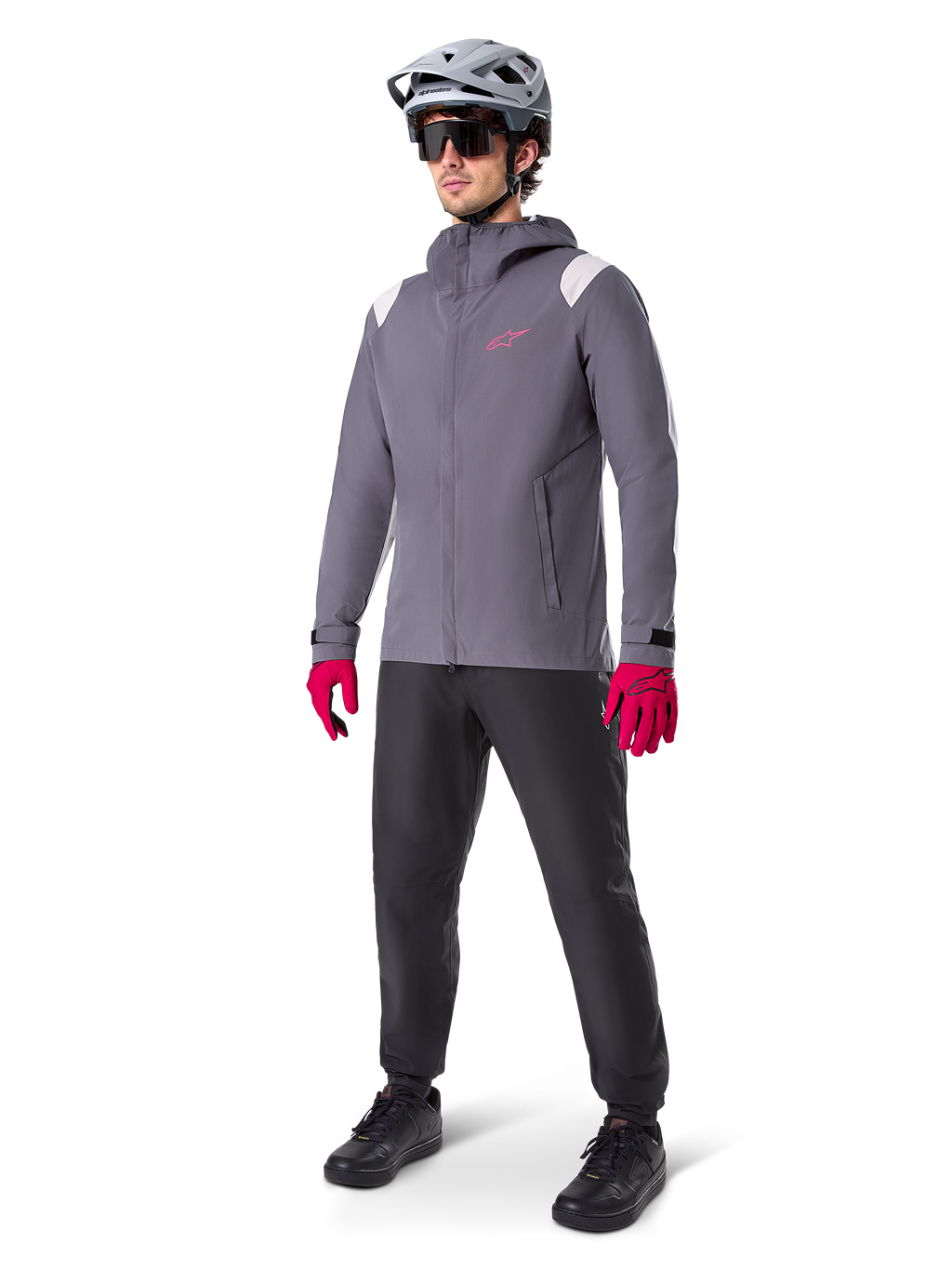 Manlig modell som bär Alpinestars A-Dura Regnjacka i Dark Titanium, grå jacka med huva med rosa logotyp på bröstet och ljusgrå axelpaneler, matchad med en grå MTB-hjälm, mörka solglasögon, starkt rosa handskar och svarta byxor för terrängcykling.