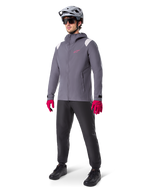 Manlig modell som bär Alpinestars A-Dura Regnjacka i Dark Titanium, grå jacka med huva med rosa logotyp på bröstet och ljusgrå axelpaneler, matchad med en grå MTB-hjälm, mörka solglasögon, starkt rosa handskar och svarta byxor för terrängcykling.