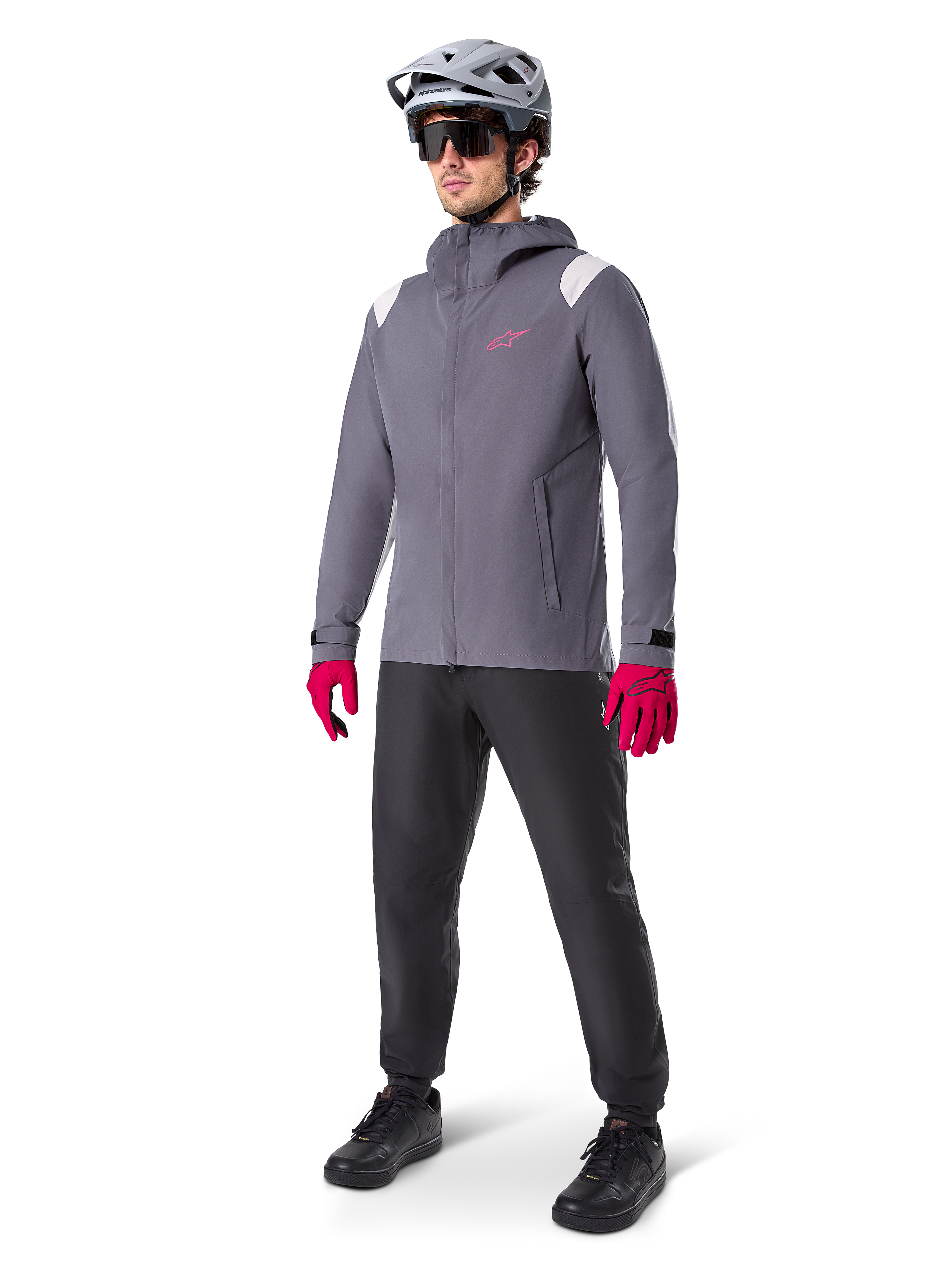 Manlig modell som bär Alpinestars A-Dura Regnjacka i Dark Titanium, grå jacka med huva med rosa logotyp på bröstet och ljusgrå axelpaneler, matchad med en grå MTB-hjälm, mörka solglasögon, starkt rosa handskar och svarta byxor för terrängcykling.