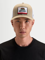Alpinestars Cali 2.0 keps, trucker-keps i khakifärg och vitt, med böjd skärm och Kalifornien-björnmärke på framsidan, bärs av en modell