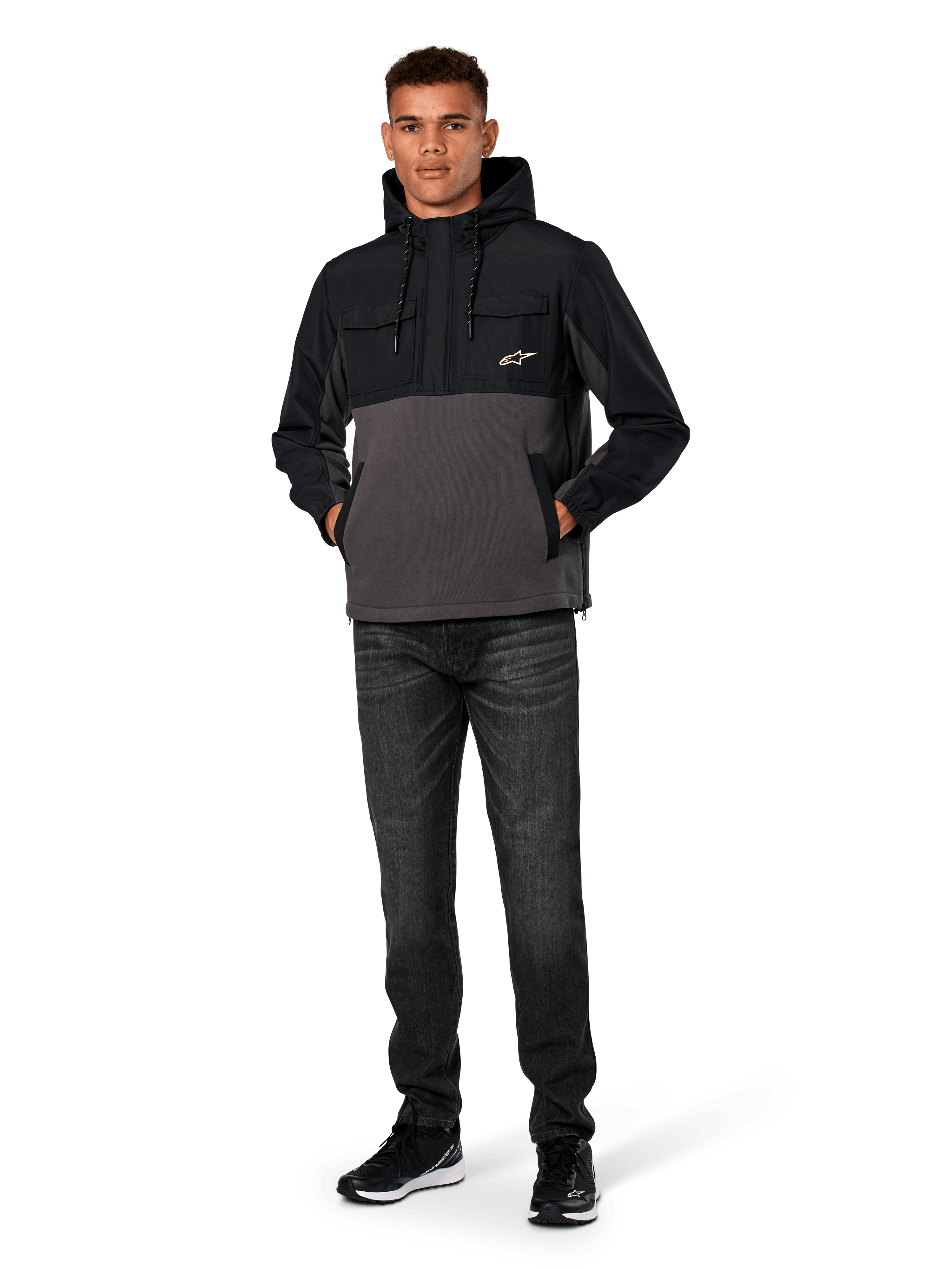 Alpinestars Juncture Hybrid-jacka, svart och gunmetal, anorak-hoodie i mixat material med kropp i canvasfleece och ripstop-överdrag, vit A-star-logotyp på bröstet, huva med dragsko, bärs av modell med mörka jeans och svarta sneakers, avslappnad motorsport-inspirerad streetwear
