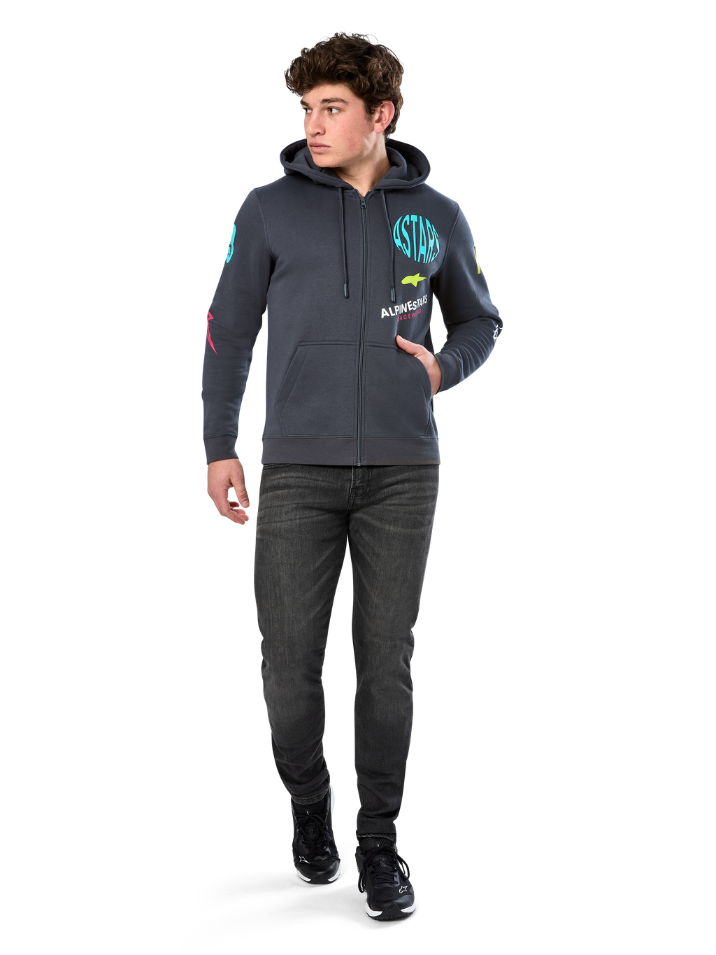 Alpinestars Variegate Hoodie, fleecehoodie med dragkedja framtill, gunmetal-grå, bärs av modell med svarta jeans och sneakers, med turkos 'ASTARS'-märkning och färgglad logografik på ärmar och bröst, avslappnad motorsportinspirerad streetwear