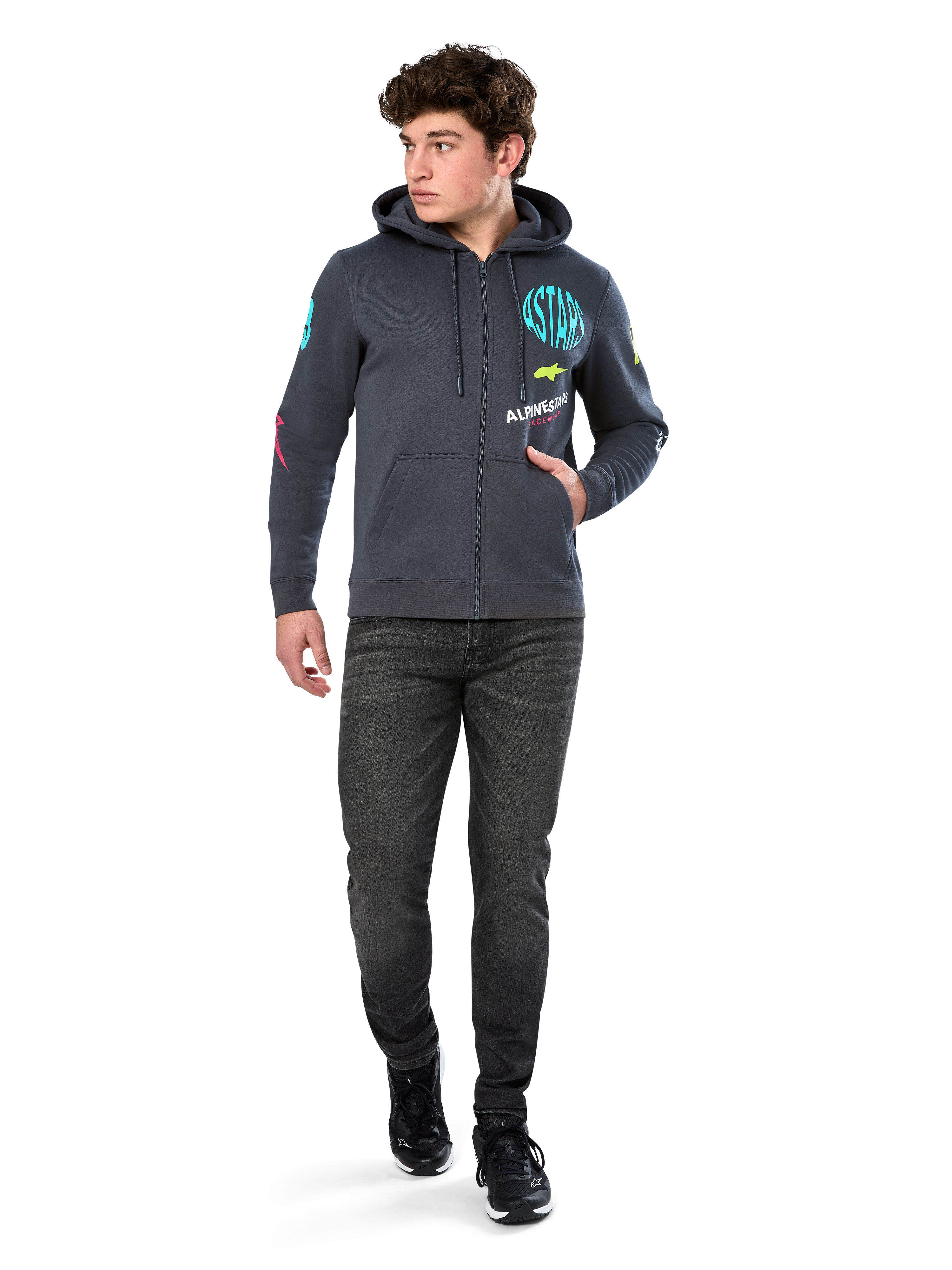Alpinestars Variegate Hoodie, fleecehoodie med dragkedja framtill, gunmetal-grå, bärs av modell med svarta jeans och sneakers, med turkos 'ASTARS'-märkning och färgglad logografik på ärmar och bröst, avslappnad motorsportinspirerad streetwear