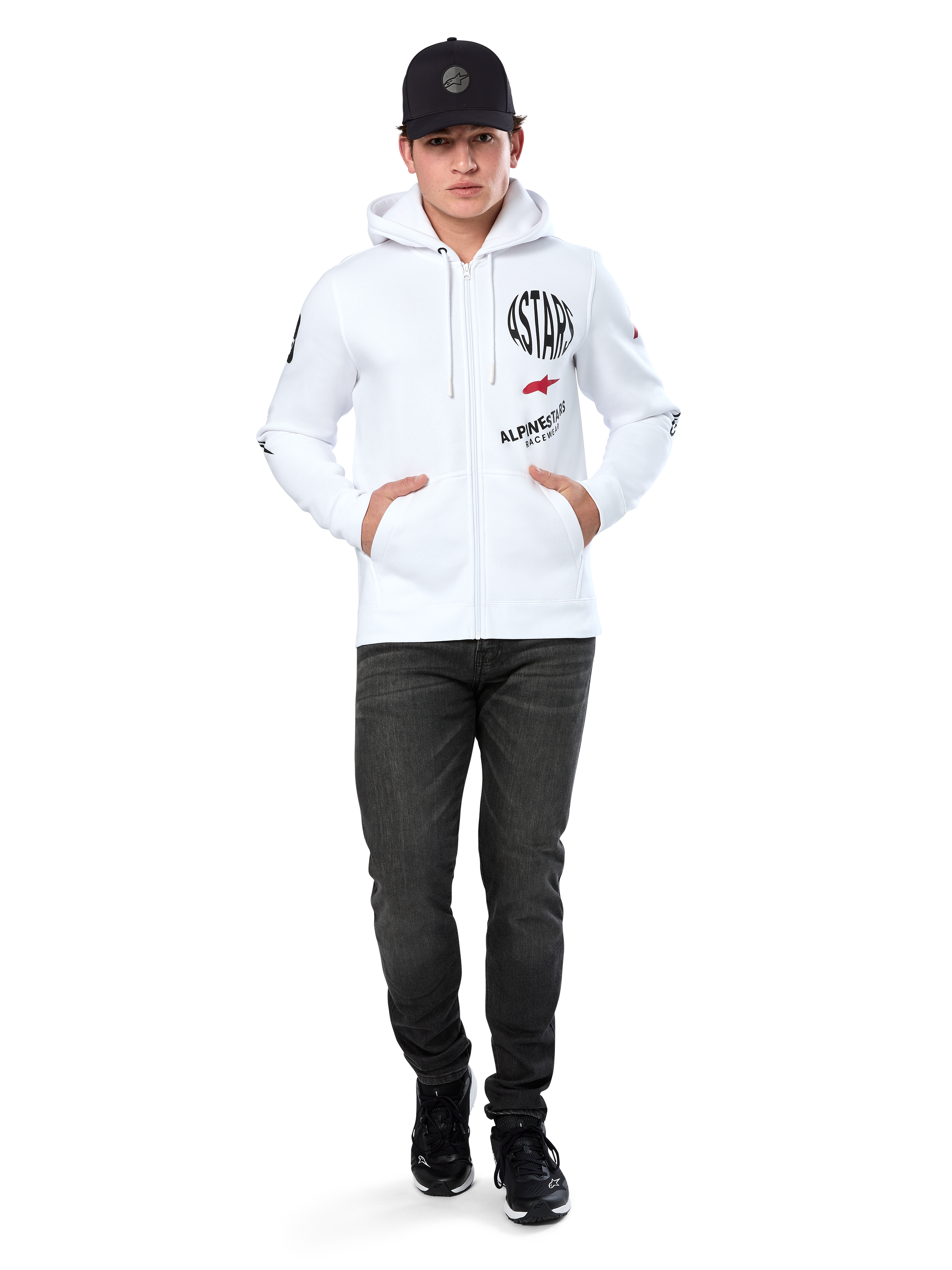 Alpinestars Variegate Hoodie, vit fleecehoodie med dragkedja, grafik med flera logotyper inklusive 'ASTARS' och röd stjärnlogotyp på bröstet, grafiska detaljer på ärmarna, bärs av modell med svart keps och svarta jeans, avslappnat motorsport-streetwear