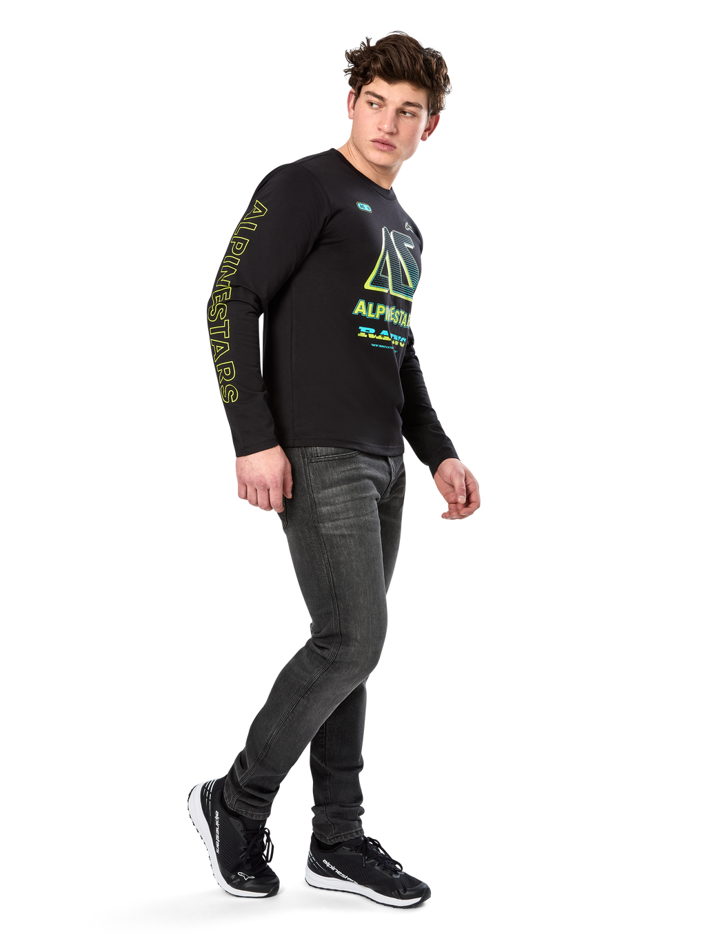 Man som bär Alpinestars Ayess CSF långärmad t-shirt, svart, med gul text på ärmarna och livfull grafik i lime och cyan på bröstet, kombinerat med mörkgrå jeans och svarta sneakers, avslappnad motorsportinspirerad streetwear.