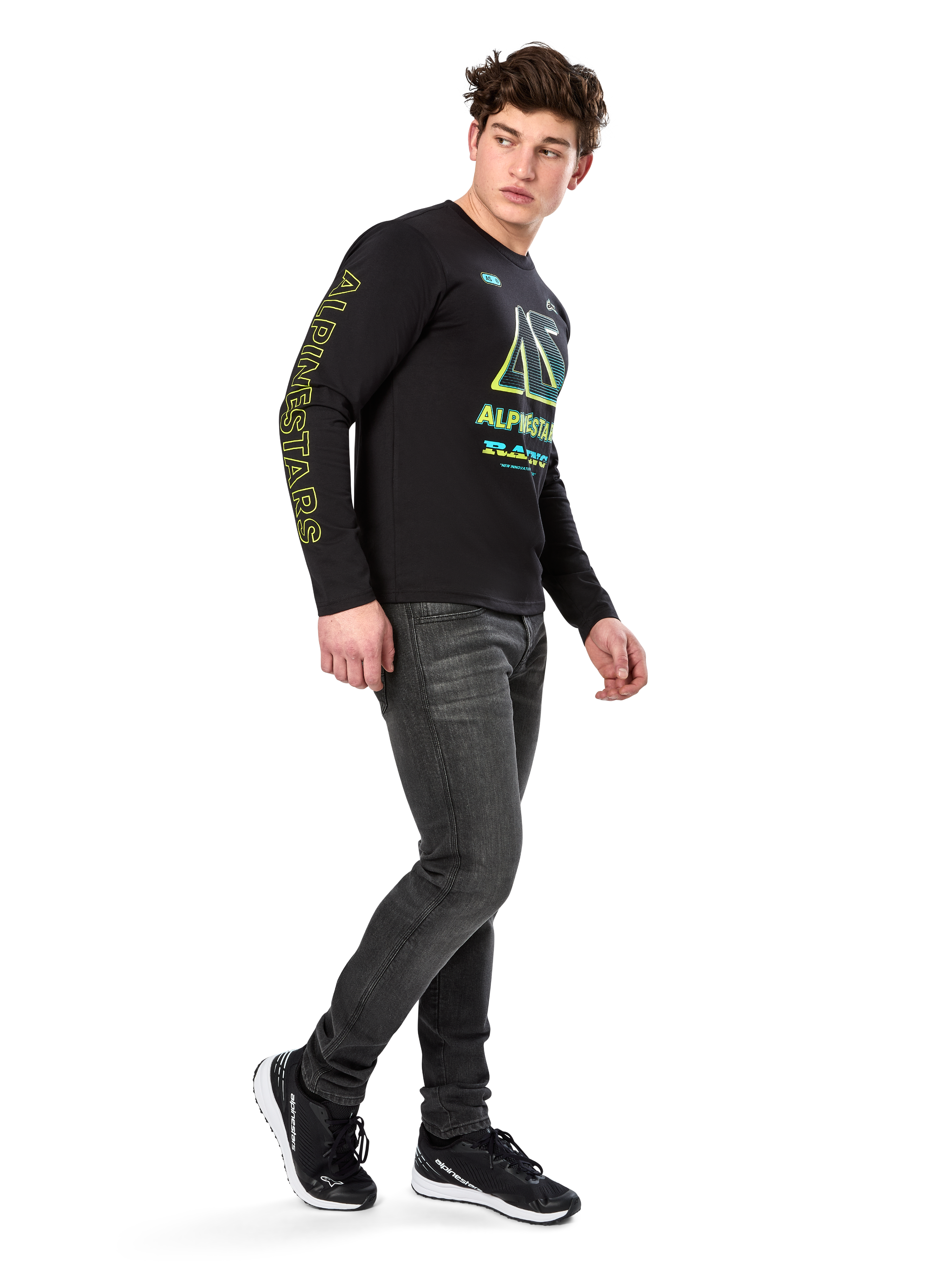 Man som bär Alpinestars Ayess CSF långärmad t-shirt, svart, med gul text på ärmarna och livfull grafik i lime och cyan på bröstet, kombinerat med mörkgrå jeans och svarta sneakers, avslappnad motorsportinspirerad streetwear.