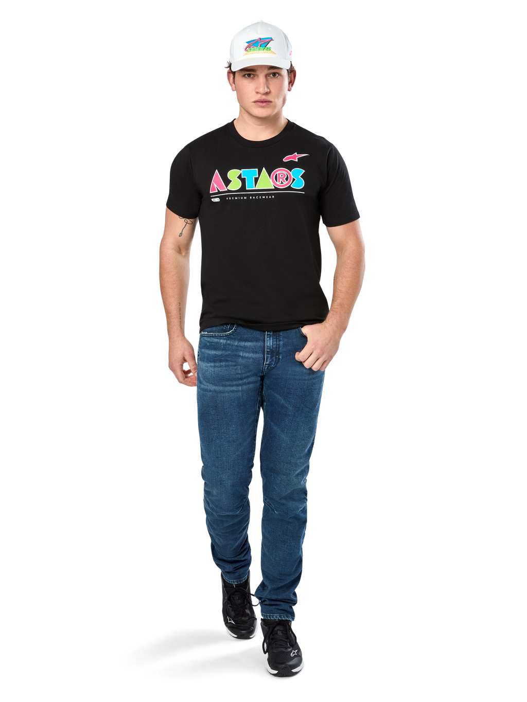 Manlig modell i Alpinestars Cereal CSF Tee, svart kortärmad t-shirt med färgglad geometrisk ASTARS-logotyp, matchad med blå jeans och vit keps, avslappnad motorsportinspirerad streetwear.