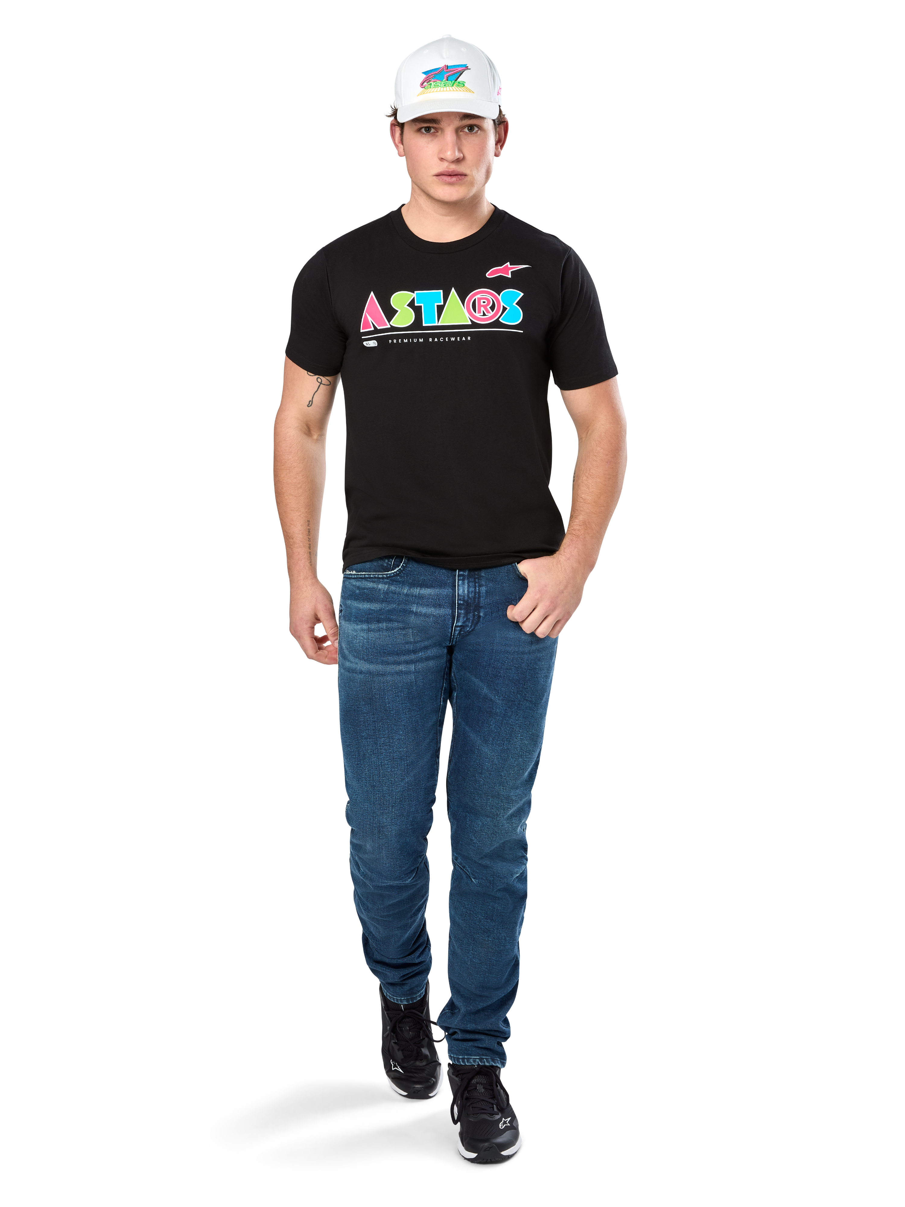Manlig modell i Alpinestars Cereal CSF Tee, svart kortärmad t-shirt med färgglad geometrisk ASTARS-logotyp, matchad med blå jeans och vit keps, avslappnad motorsportinspirerad streetwear.