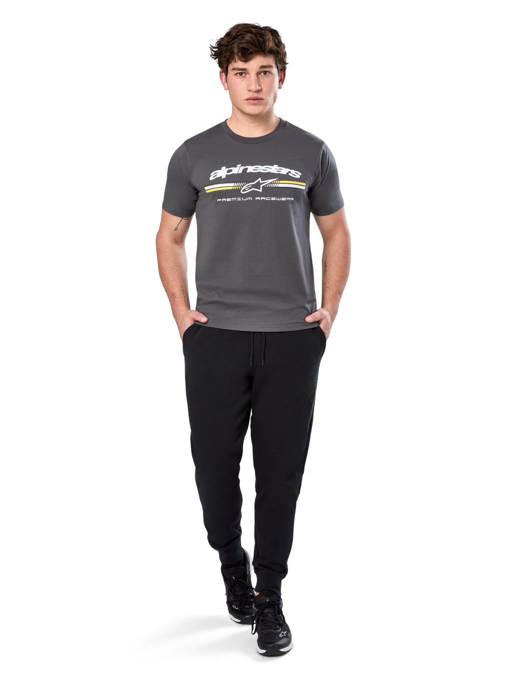 Man i Alpinestars Prevail Csf Tee, kortärmad t-shirt, mörkgrå, med vit Alpinestars-logotyp och trefärgad rand med gula detaljer, matchad med svarta joggers och sneakers, avslappnad motorsport-streetwear