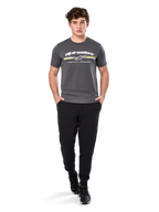 Man i Alpinestars Prevail Csf Tee, kortärmad t-shirt, mörkgrå, med vit Alpinestars-logotyp och trefärgad rand med gula detaljer, matchad med svarta joggers och sneakers, avslappnad motorsport-streetwear