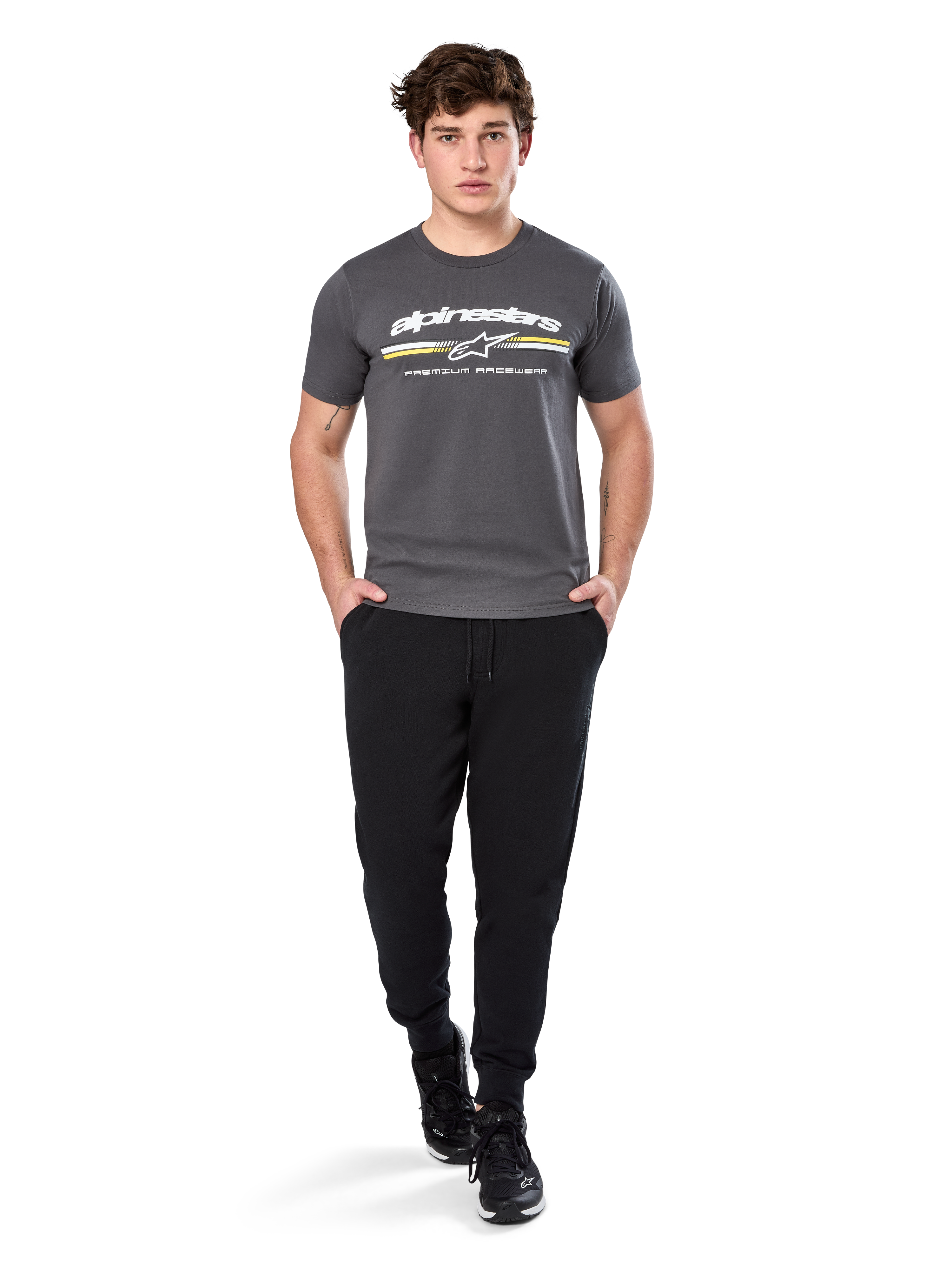 Man i Alpinestars Prevail Csf Tee, kortärmad t-shirt, mörkgrå, med vit Alpinestars-logotyp och trefärgad rand med gula detaljer, matchad med svarta joggers och sneakers, avslappnad motorsport-streetwear