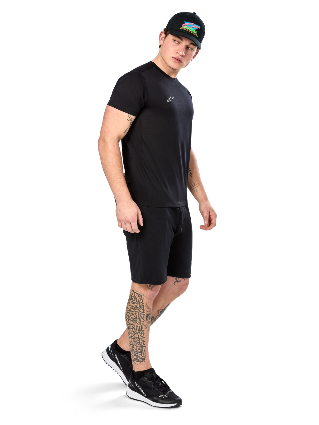 Man med tatueringar bär Alpinestars Point Performance Tee, kortärmad svart funktions-t-shirt med liten vit logotyp, matchad med svarta shorts, en svart keps med färgglad grafik och svartvita sneakers, sportig casual livsstil