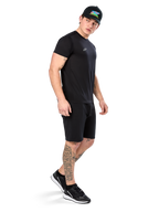 Man med tatueringar bär Alpinestars Point Performance Tee, kortärmad svart funktions-t-shirt med liten vit logotyp, matchad med svarta shorts, en svart keps med färgglad grafik och svartvita sneakers, sportig casual livsstil