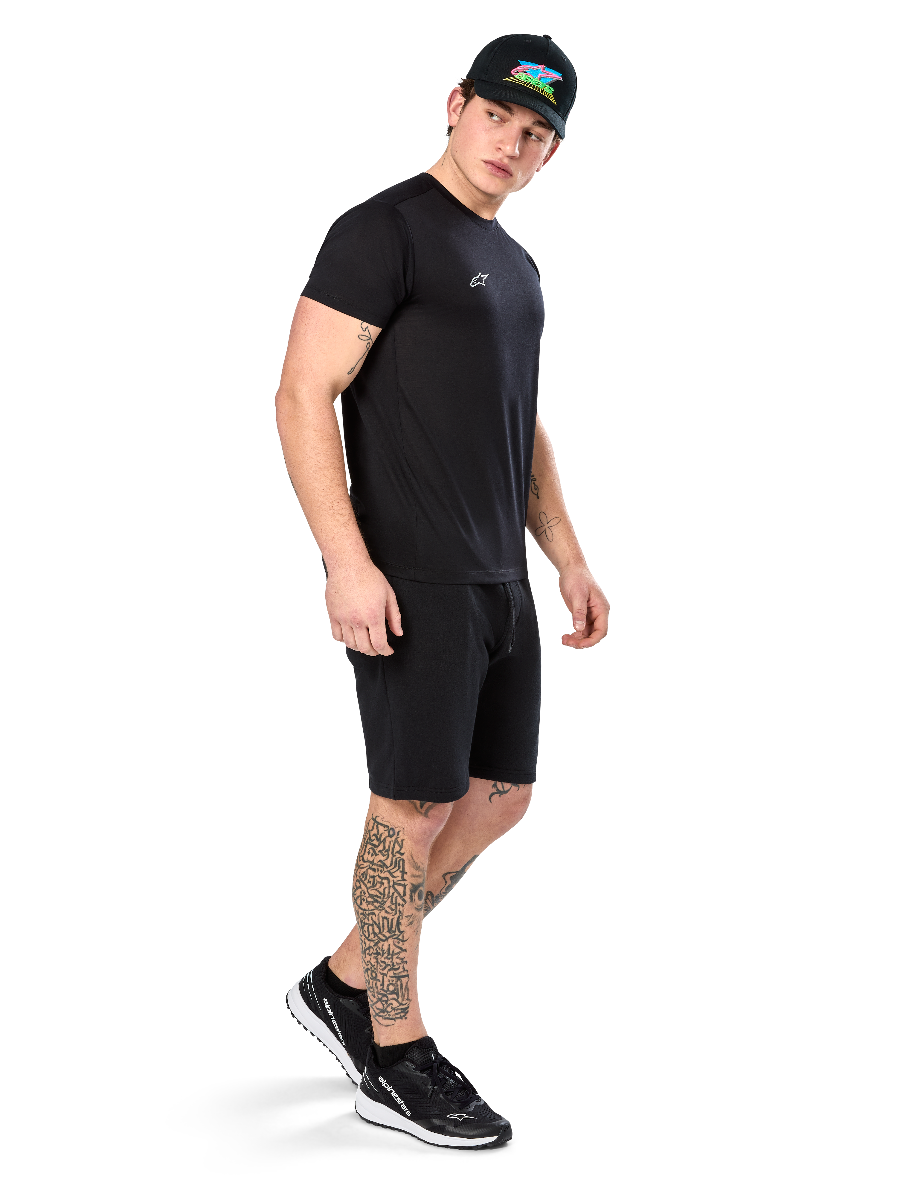 Man med tatueringar bär Alpinestars Point Performance Tee, kortärmad svart funktions-t-shirt med liten vit logotyp, matchad med svarta shorts, en svart keps med färgglad grafik och svartvita sneakers, sportig casual livsstil