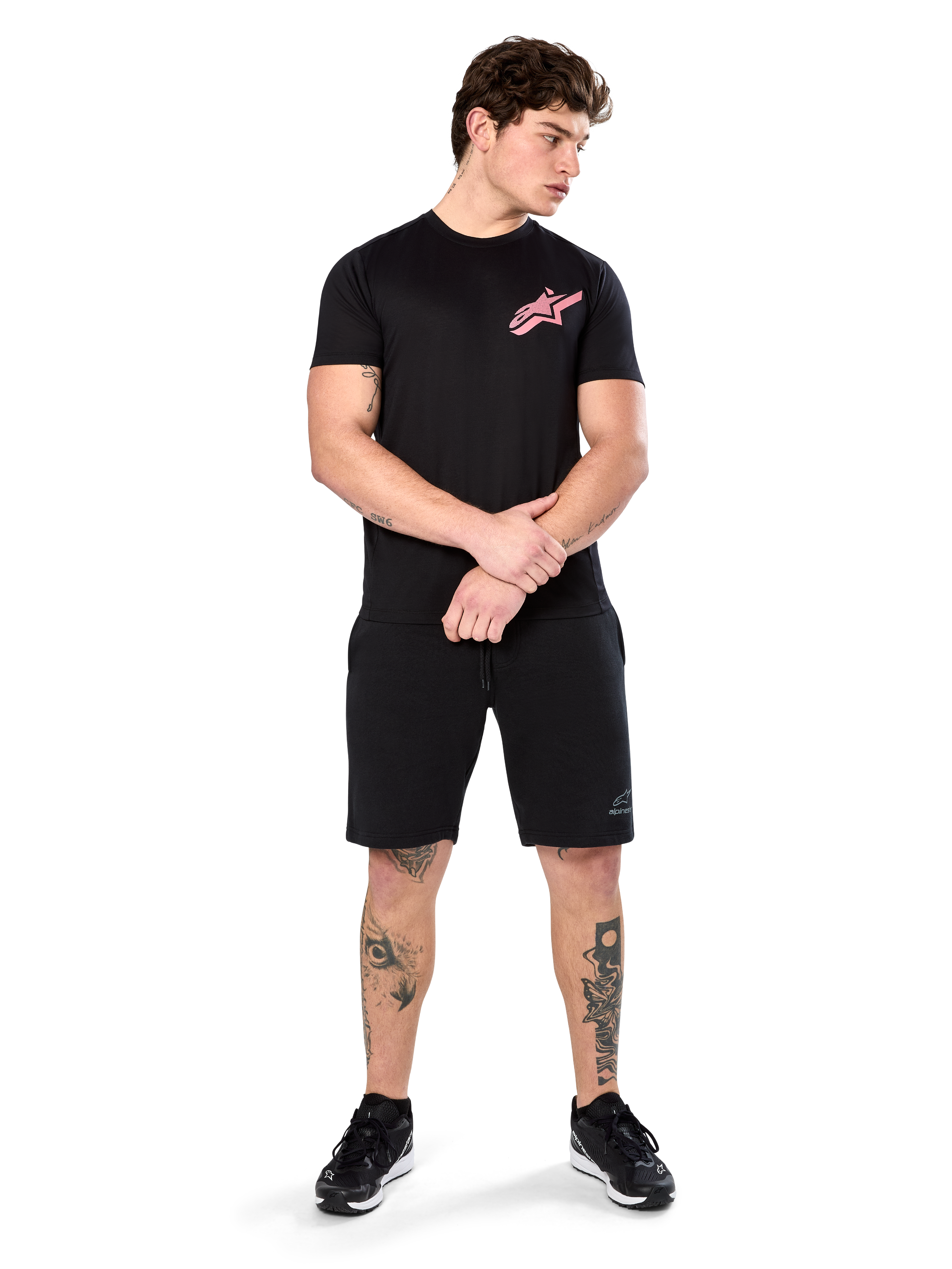 Model wearing Alpinestars Attrition Performance Tee, Short Sleeve Performance T-Shirt, Black with a pink Astars logo on the chest, paired with black shorts and sneakers, casual motorsport-inspired sportswear