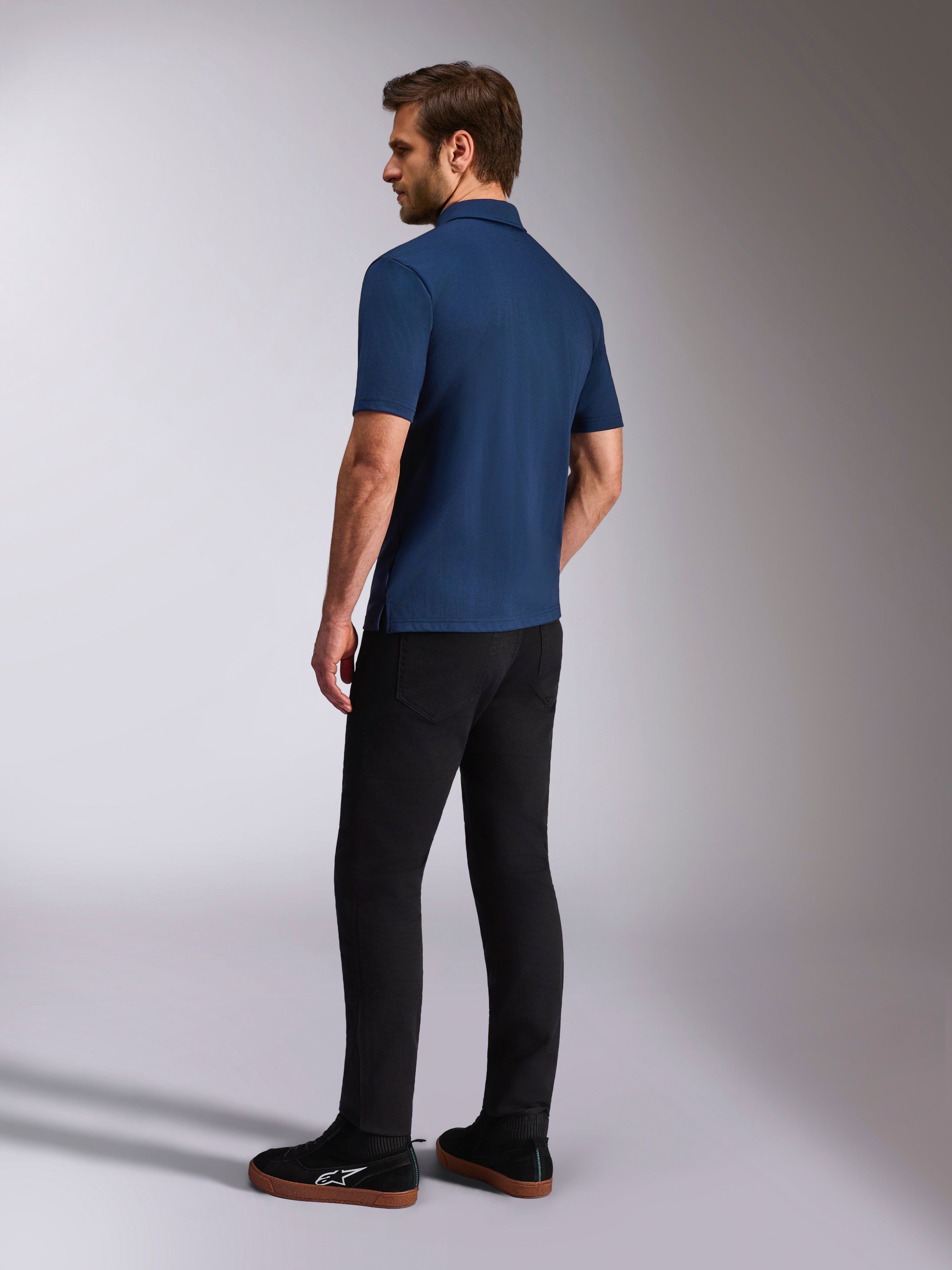 Alpinestars Impetus Sport pikétröja, marinblå kortärmad pikétröja, bak- och sidovy på modell, kombinerad med svarta byxor och svarta sneakers, avslappnade sportkläder för en motorsport-livsstil