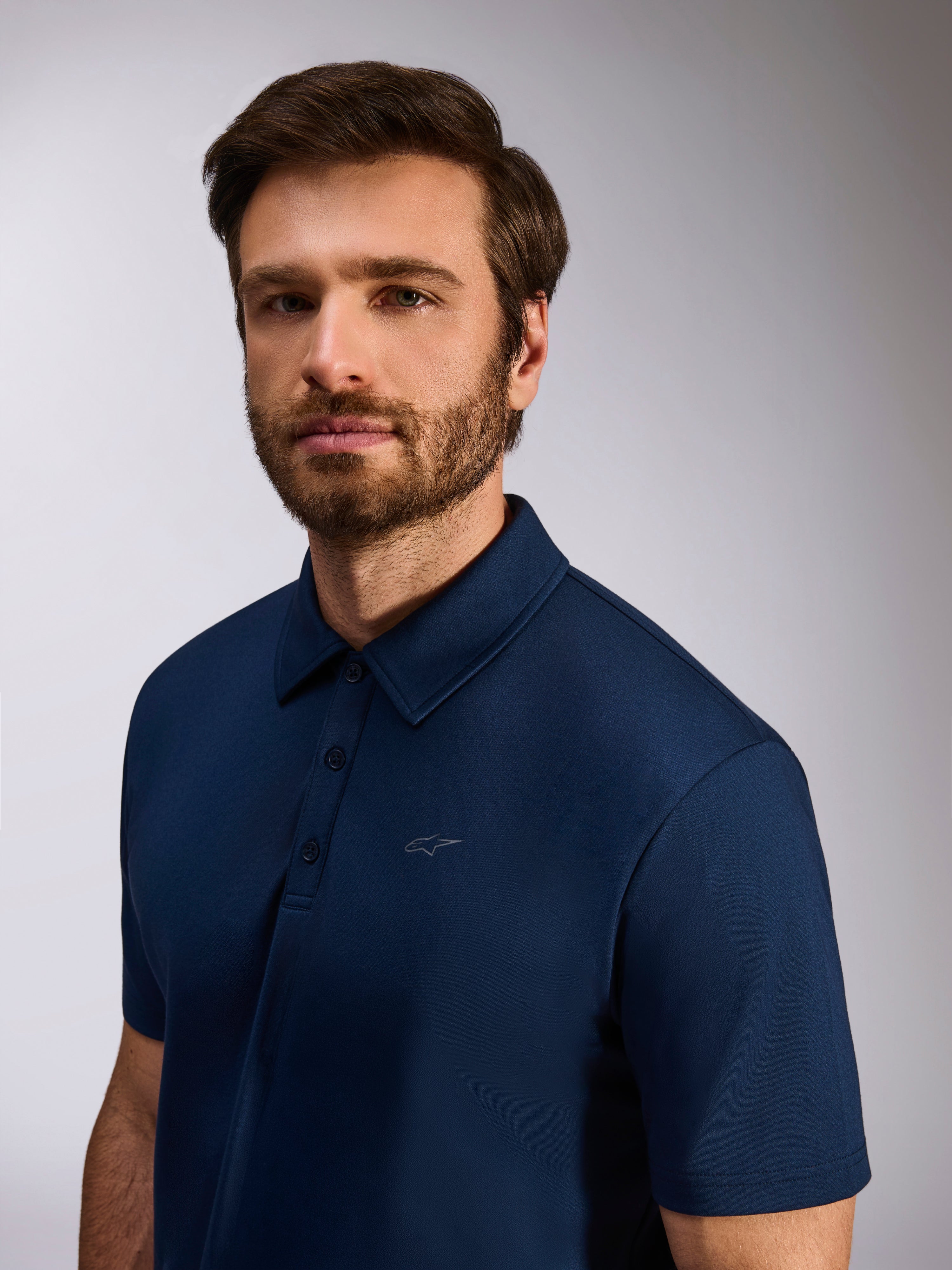 Modell bär Alpinestars Impetus Sport Polo, marinblå, kortärmad pikétröja i mock mesh, med tre knappar och en subtil Astars-logotyp på bröstet, casual livsstilskläder för motorsport